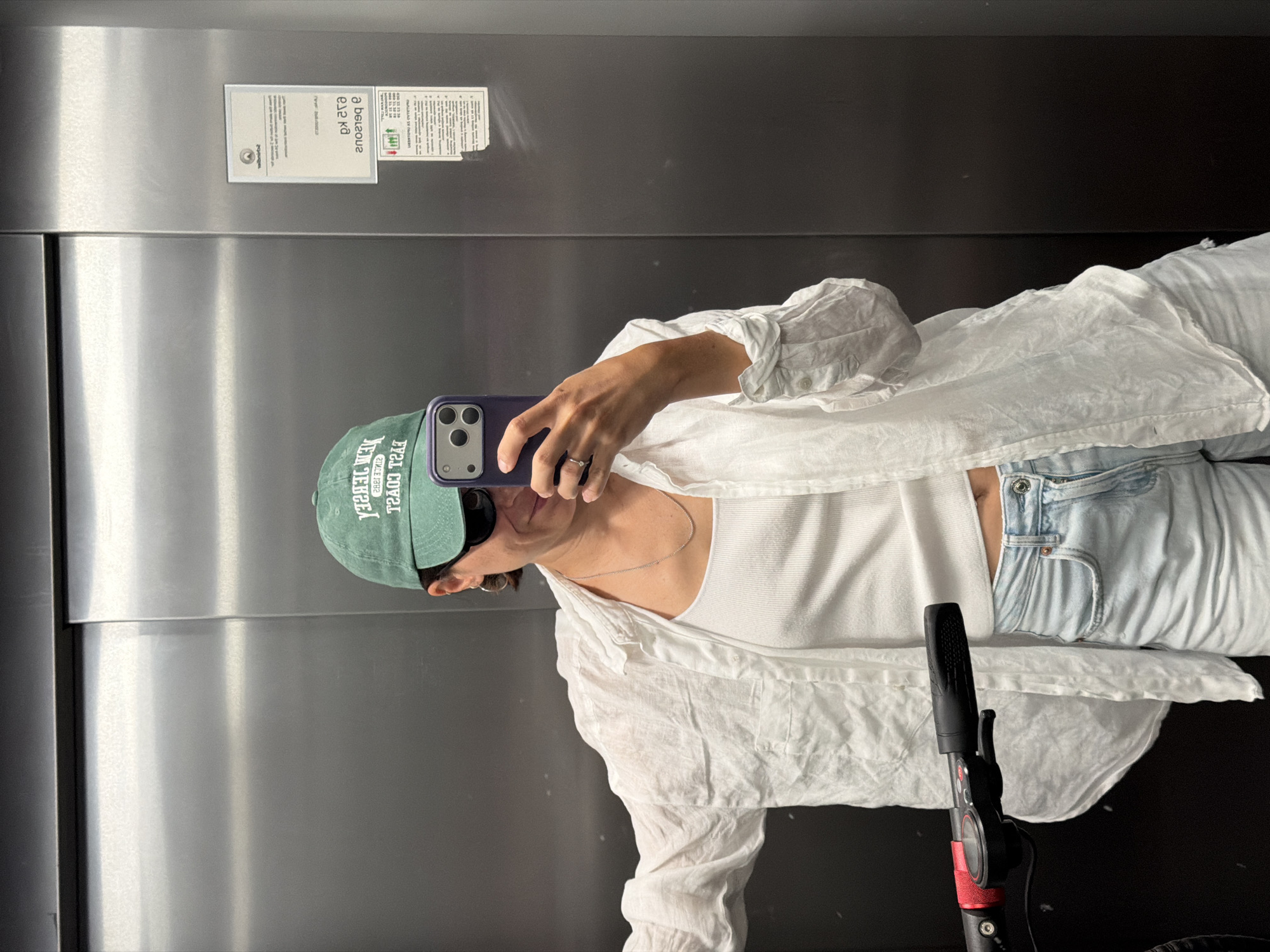 A person is taking a selfie in an elevator with stainless steel doors. The person is wearing a green baseball cap with white lettering, sunglasses, a white tank top, a white linen shirt, and light blue jeans. They are holding a purple iPhone in their right hand. A silver chain necklace is visible around their neck, as well as a ring on their finger. The handlebars of an electric scooter are partially visible in the lower center of the frame. Two signs are adhered to the elevator door. The location is speculated to be in Budva, Montenegro.