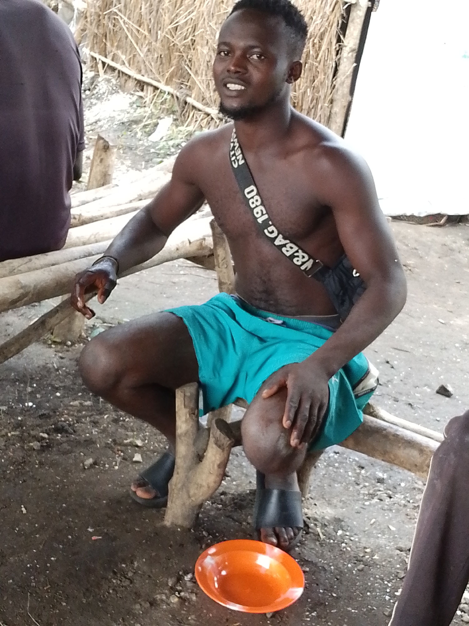 A shirtless young Black man with dark skin and short, dark hair is seated on a rustic wooden bench in what appears to be an outdoor setting. He is wearing teal-colored shorts and black slides. A dark strap with white lettering crosses his chest. He is looking towards the viewer with a slight smile and his head is tilted. In front of him, on the ground, is a bright orange plate. The background consists of dried grass or straw and some wooden structures. To the left of the man, another person's back is visible, dressed in a dark shirt. The ground is dirt with scattered small stones and debris. The lighting suggests it is daytime, possibly outdoors in a somewhat shaded area. The scene has a casual, possibly rural or informal atmosphere. Visible text on the strap reads "CITY NEWS VBAG. 1980".