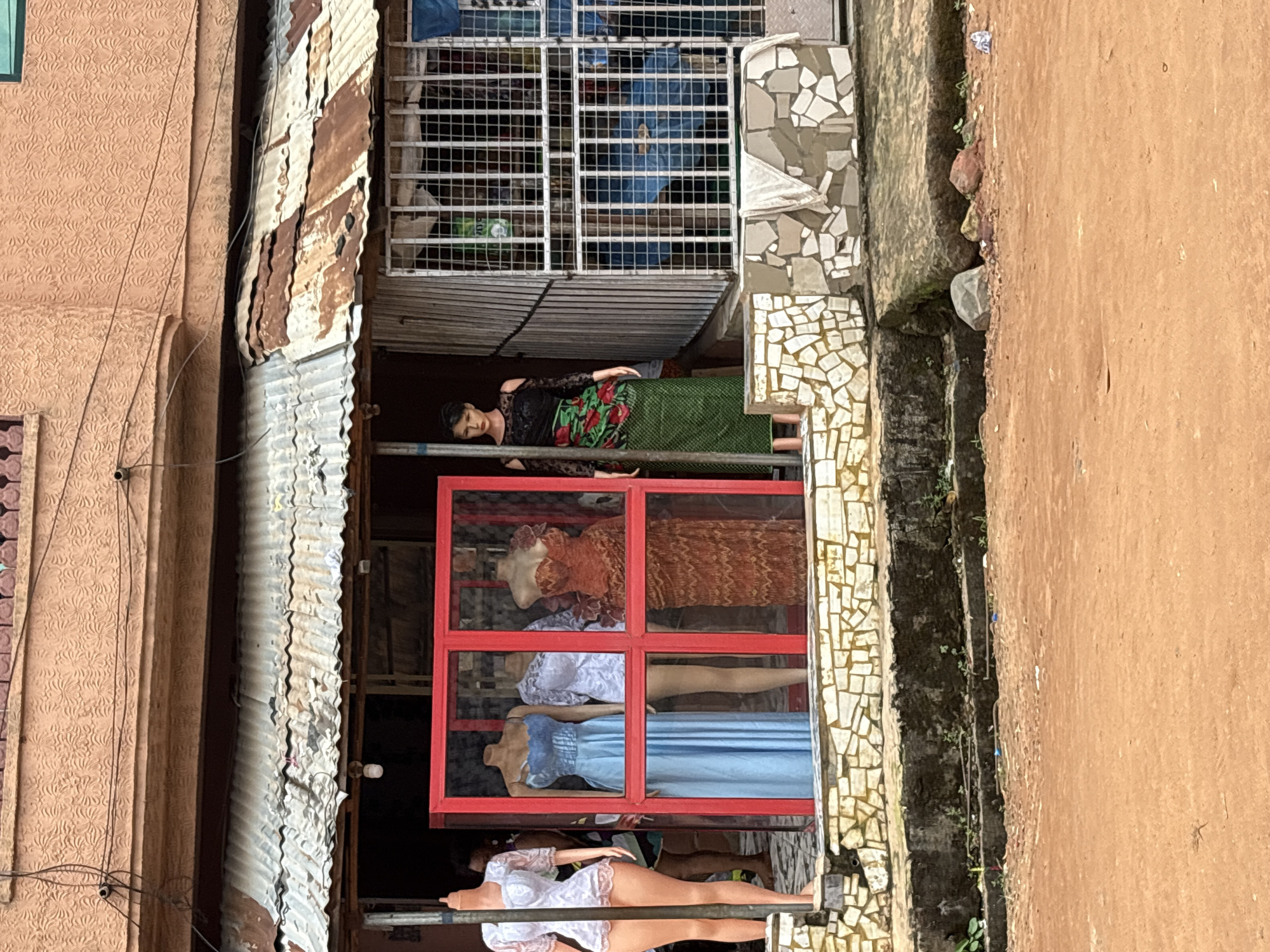 This image depicts a street scene in Onitsha, Nigeria, likely a commercial area given the presence of a shop displaying mannequins and clothing. The shop, situated at street level, has a corrugated metal roof that appears weathered and rusted. The facade is decorated with a mosaic of white and beige tiles.
Inside a red-framed display area, three mannequins are visible. The leftmost mannequin is headless and wears a white lace garment. To its right, a mannequin in a light blue dress stands next to a headless mannequin in a white lace dress. The rightmost mannequin in this section wears an orange, intricately patterned dress. To the right of this red-framed display, a fourth mannequin stands with its head, wearing a black top with off-the-shoulder sleeves and a vibrant green and red patterned skirt. Behind the mannequins, glimpses of the shop's interior reveal stored items, including what appears to be blue fabric.
The shop's entrance is protected by a metal security grate. The ground in front of the shop is rough, with a concrete step leading up to the storefront, also adorned with the mosaic tile pattern. The area directly in front of the shop is covered with reddish-brown earth, with some debris and scattered rocks visible. The overall impression is of a bustling street market environment.