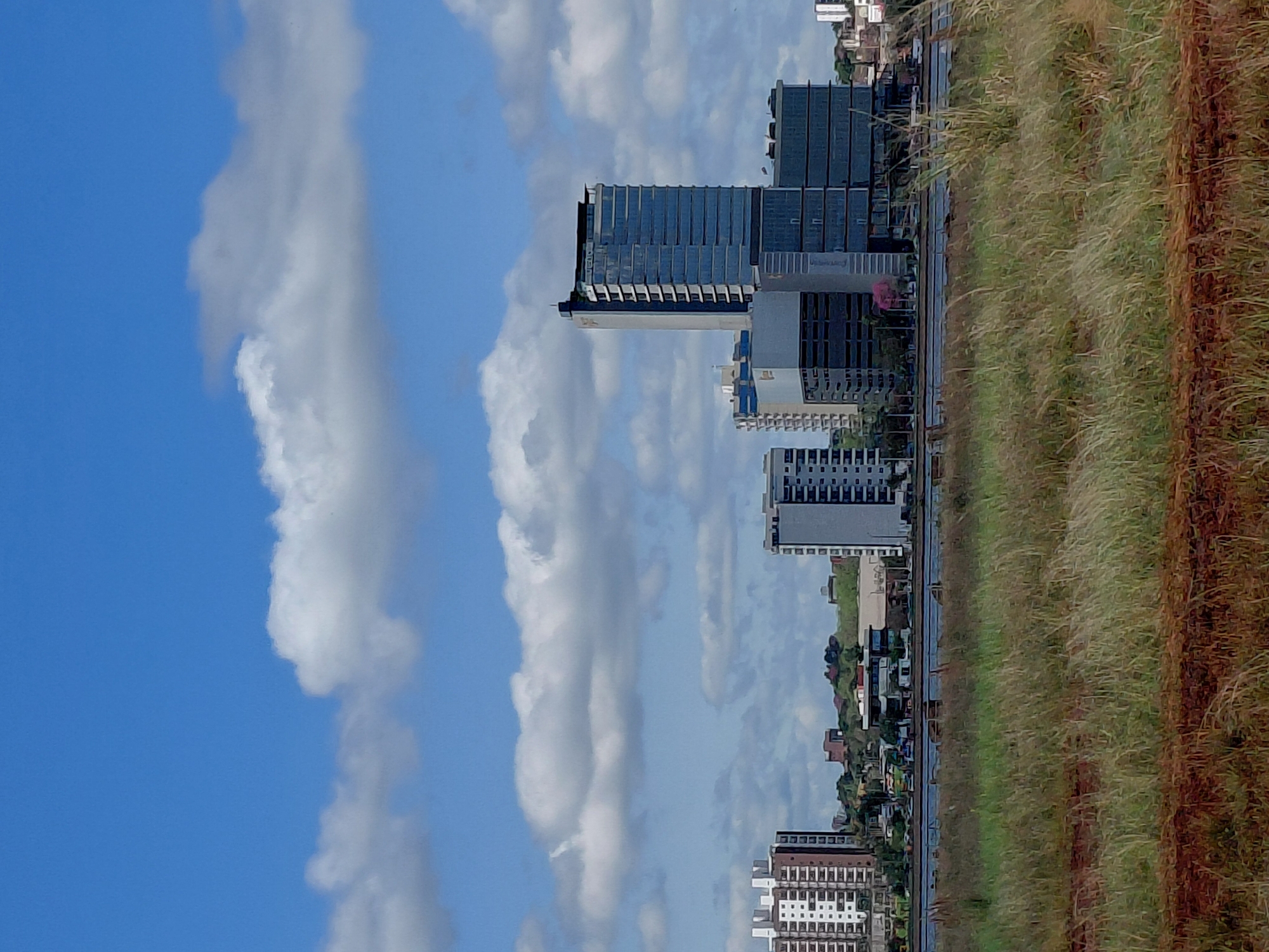 The image depicts a modern cityscape in Encarnación, Paraguay, characterized by contemporary architecture and a vibrant blue sky dotted with fluffy white clouds. In the foreground, a grassy field stretches across the frame, with a strip of reddish-brown earth bordering its lower edge. Beyond the field, a body of water, likely the Paraná River, separates the foreground from the urban landscape.
Dominating the middle ground are several multi-story buildings, showcasing a mix of glass facades and concrete structures. A tall, slender skyscraper with a predominantly glass exterior stands out amongst the other buildings, its reflective surfaces mirroring the sky. To its left, a slightly shorter building with a similar glass facade is visible, while further to the left, another building with a white facade and numerous windows is present. The buildings are nestled amongst a backdrop of rolling green hills and some smaller structures, suggesting a developing area within the city. The overall impression is one of urban development and modern infrastructure set against a natural landscape.