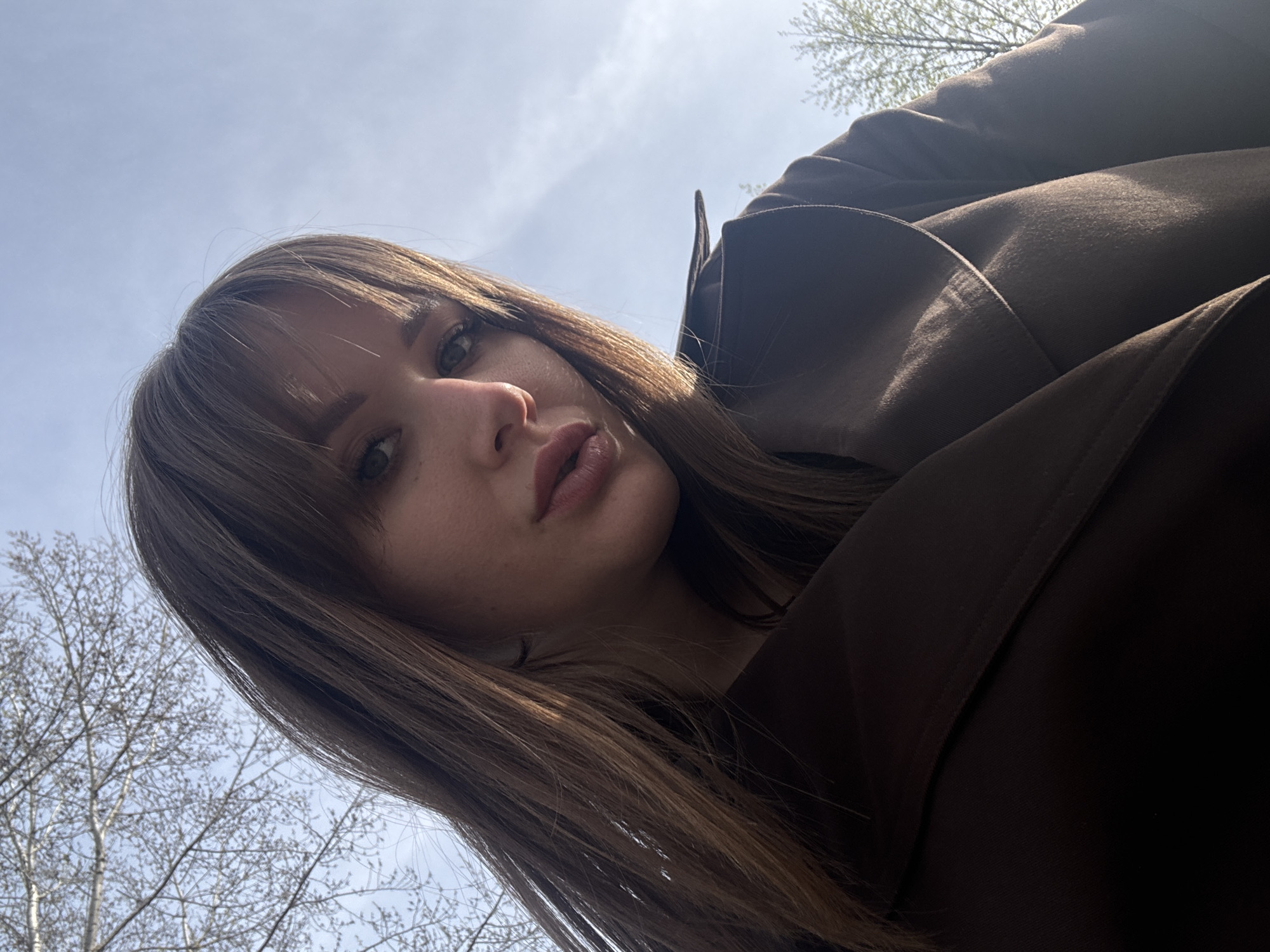 A low-angle, medium shot of a woman outdoors on a partly cloudy day. The woman has shoulder-length, light brown hair with bangs, and is wearing a brown jacket or coat. Bare trees with sparse foliage are visible in the background against the sky. The location is likely the city of Chaikovsky, Russia.