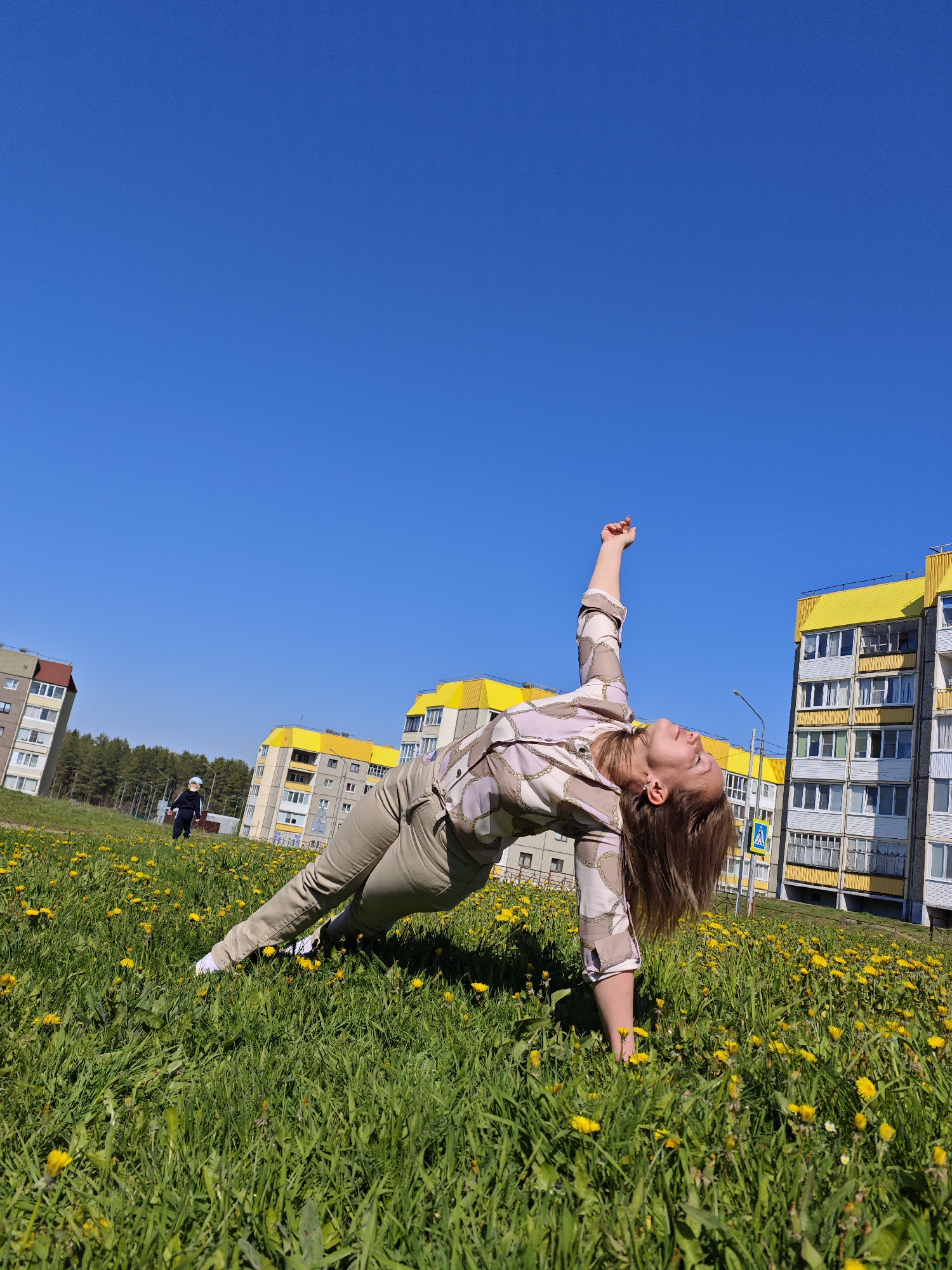 A full shot captures a woman performing a yoga pose on a grassy field filled with dandelions, under a clear blue sky in Марковский, Russia. The woman is positioned diagonally across the frame, with her weight supported by one arm and leg, and her other arm raised towards the sky. She is wearing beige pants and a patterned long-sleeved shirt. In the background are multi-story residential buildings with yellow roofs and grey facades. To the left, a child walks on the field. A street lamp and a pedestrian crossing sign are also visible in the background.