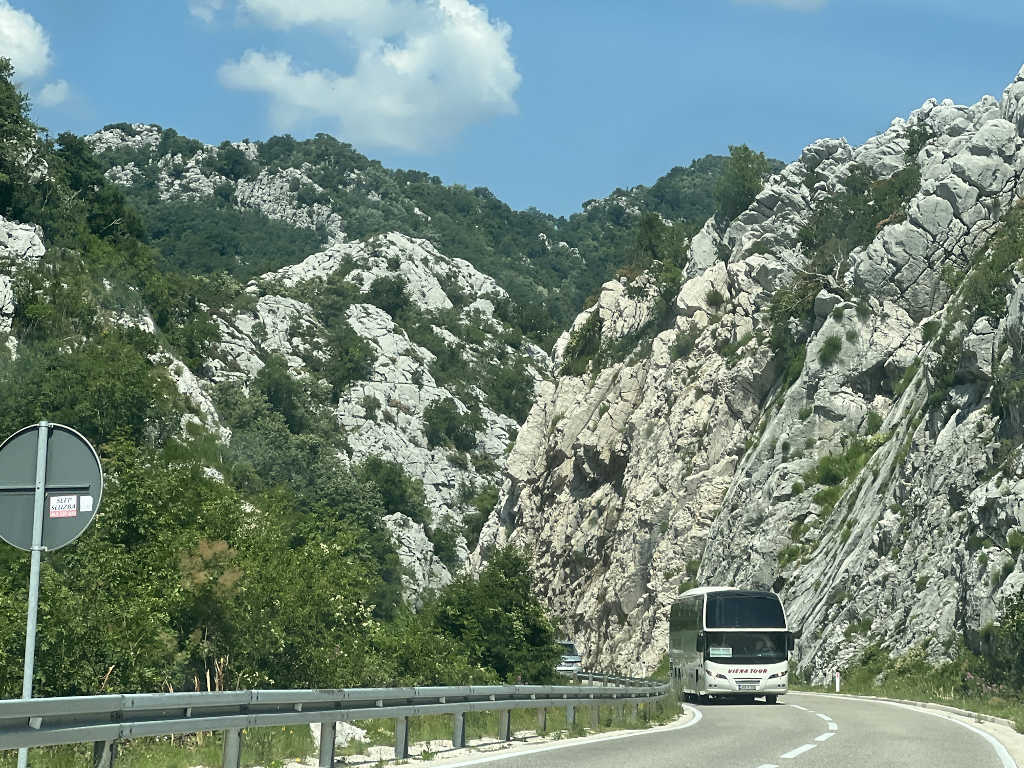 The image depicts a scenic mountain road in the vicinity of Dragalj, Montenegro. A white tour bus with "VIENA TOUR" branding is traveling on the winding asphalt road, which is bordered by a metal guardrail. To the left of the road, a circular sign with a cross symbol and a sticker reading "SLÈP SLUŽBA" is visible. The surrounding environment is characterized by steep, rocky mountainsides covered with dense green vegetation. The sky above is clear blue with scattered white clouds. The overall scene conveys a sense of travel and exploration through a rugged natural landscape.