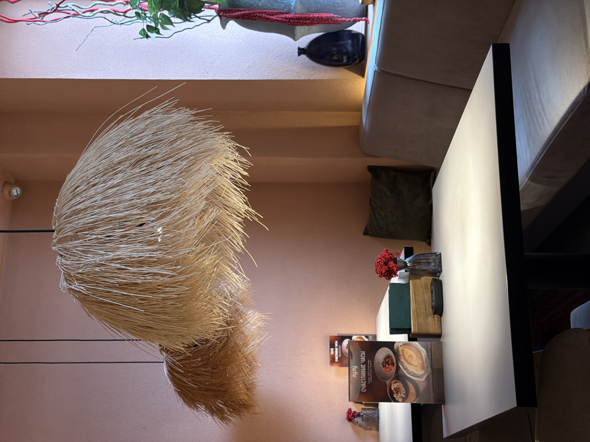 This image depicts the cozy interior of a cafe or restaurant located in Chaikovsky, Russia, characterized by warm, artificial lighting. The scene is set up for dining or relaxation but is currently devoid of people.
In the foreground, a striking, large decorative light fixture hangs, composed of numerous thin, light-colored strands resembling straw or raffia, fanned out to form a voluminous, spherical shape. A lighter-toned wall, possibly painted in a muted pink or beige, serves as the background, featuring a small, circular light fixture.
To the right, a comfortable dining area is arranged with a plush, light-colored upholstered booth or bench seat, accented by a dark green cushion. Next to the seating is a clean, rectangular table with a light-colored surface and dark edges. On the table, various items are neatly placed: a clear glass vase holding vibrant red berry-like flowers, a small wooden block, a folded green napkin, and a dark circular object.
Above the seating area, a narrow shelf or ledge showcases decorative elements, including green leafy plants intertwined with reddish-pink and turquoise branches, alongside a dark-colored vase.
Several menus are visible on the table. One prominently displays text in Cyrillic: "СЧАСТЛИВЫЕ ЧАСЫ" (Happy Hours) and "ЛУЛУ" (Lulu). Smaller text details a "скидка 20% на всё меню" (20% discount on the entire menu) available "В будние дни с 12 до 16" (on weekdays from 12 to 16). The overall atmosphere suggests an inviting and comfortable spot for patrons.