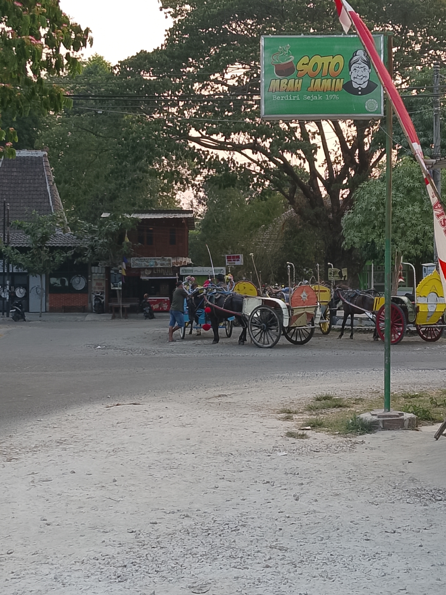 The image captures a street scene in Balun, Indonesia, likely during the late afternoon or early evening, as indicated by the soft, diffused light and the silhouetted trees. In the foreground, a sandy, graveled surface transitions into a paved road. Several horse-drawn carriages, known locally as "andong" or "cidomo," are parked along the roadside, some with colorful decorations. One man, dressed in a green t-shirt and blue shorts, stands near the front of a carriage, seemingly attending to the horse. Further back, more carriages are visible, some with horses harnessed and ready.
Dominating the background is a lush canopy of trees, with a large, mature tree in the center, its branches extending widely. Above the trees, a sign advertises "SOTO MBAH JAMIN" with the founding year "Berdiri Sejak 1976," suggesting a local eatery. Adjacent to the sign is a flagpole with the Indonesian flag. Several buildings line the street, appearing to be shops or eateries, with some displaying signage and various items on display. The overall atmosphere is one of a lively, everyday scene in a bustling part of Balun, likely a popular spot for both locals and tourists to experience traditional transportation.