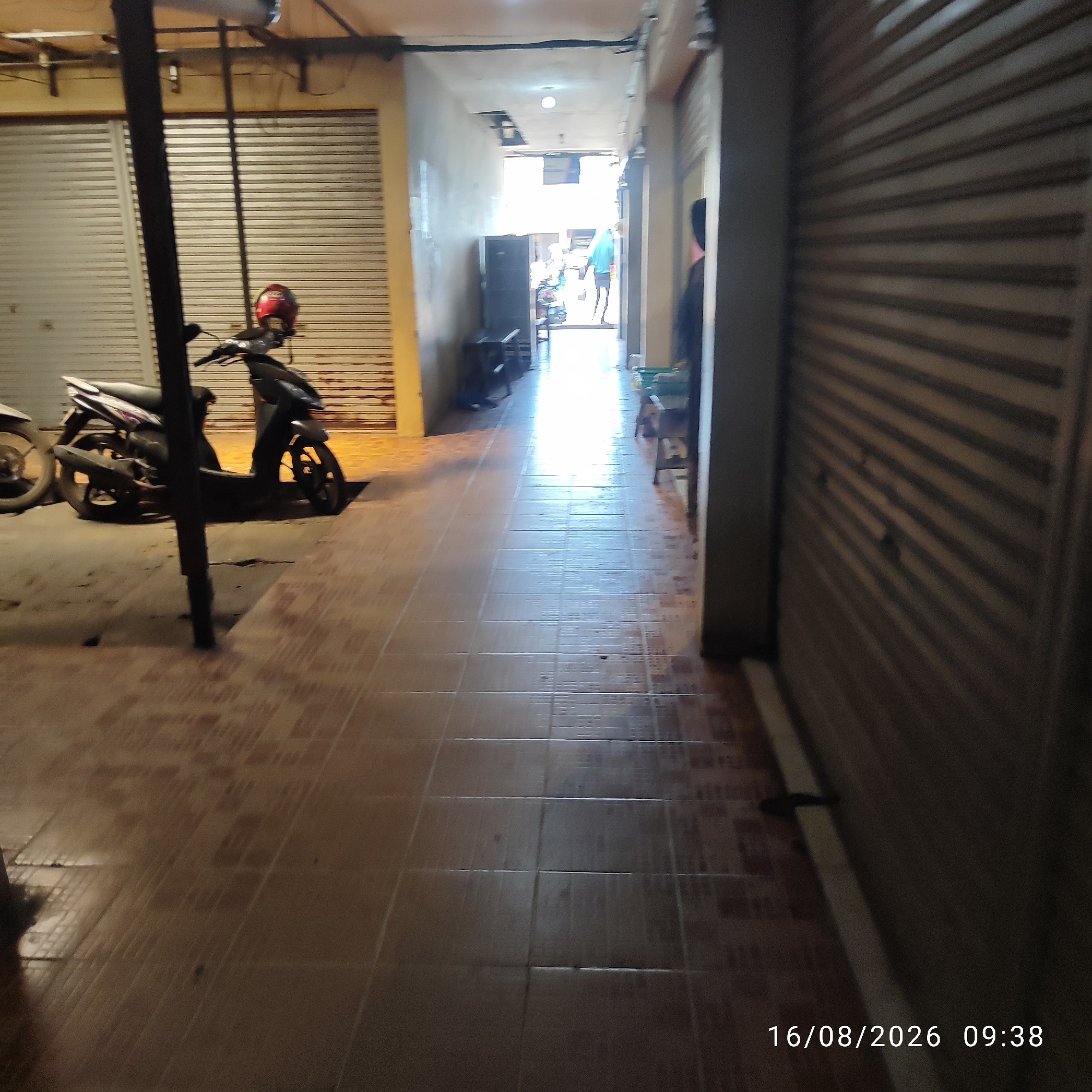The image depicts a narrow, outdoor alleyway in what appears to be a commercial area, possibly in the city of Pembaharuan, Indonesia, as indicated by the timestamp. On the left side, a dark-colored scooter with a red helmet on its handlebars is parked. The scooter is positioned in front of a closed, corrugated metal storefront. The alleyway floor is tiled with a pattern of light brown and darker brown squares. The walls of the alley are painted a light, muted color. Further down the alley, the area is brighter due to natural light entering from an opening at the end. In the distance, a person wearing a light blue garment is walking away from the camera. The right side of the alley is lined with a similarly closed, corrugated metal storefront, creating a tunnel-like effect. The overall scene suggests a quiet, possibly early morning or late afternoon atmosphere given the lighting conditions and the closed shops.