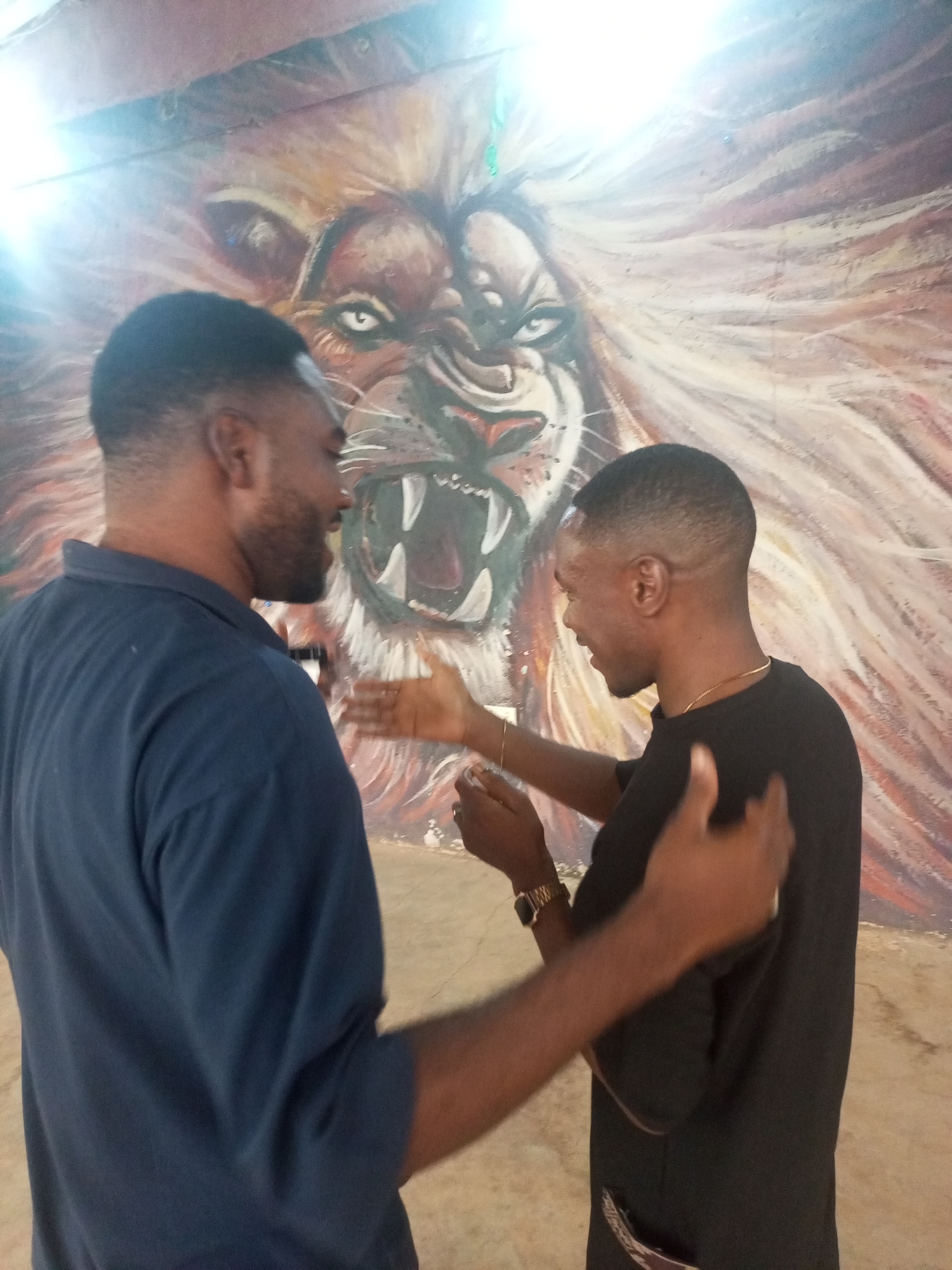 Two dark-skinned men face each other in an indoor setting, possibly Jos, Nigeria. The man on the left wears a dark blue shirt and has a short haircut with a receding hairline and a short beard. He faces right, looking at the other man. The man on the right wears a black shirt and a thin gold chain. He has a short haircut and wears a gold wristwatch on his left wrist. They both have their hands raised, appearing to interact or gesture. Behind them, a large painting of a roaring lion dominates the background. The painting depicts a fierce lion with visible teeth and a mane of light brown and white hues. The floor is a light beige color.