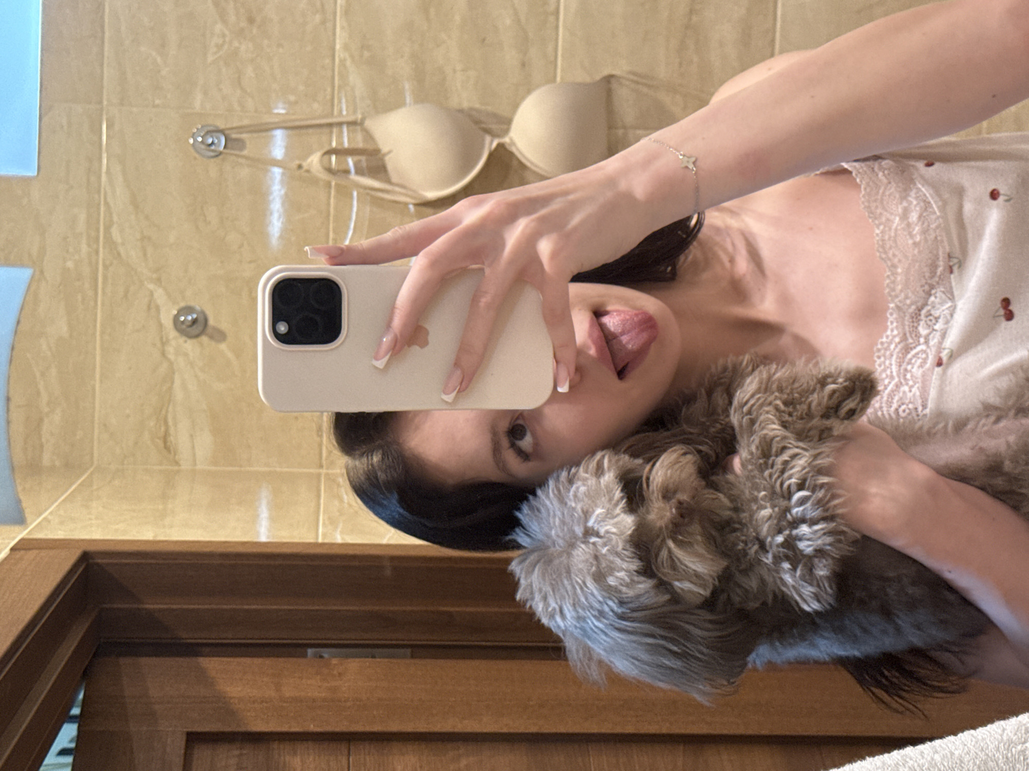 A young woman with dark hair takes a mirror selfie in what appears to be a bathroom in Khimki, Russia. She is holding a small, fluffy brown dog and sticking her tongue out. The woman is wearing a white camisole with a cherry print and lace trim. A beige bra is hanging on hooks on the tiled wall behind her. A wooden door is visible to the left of the frame.