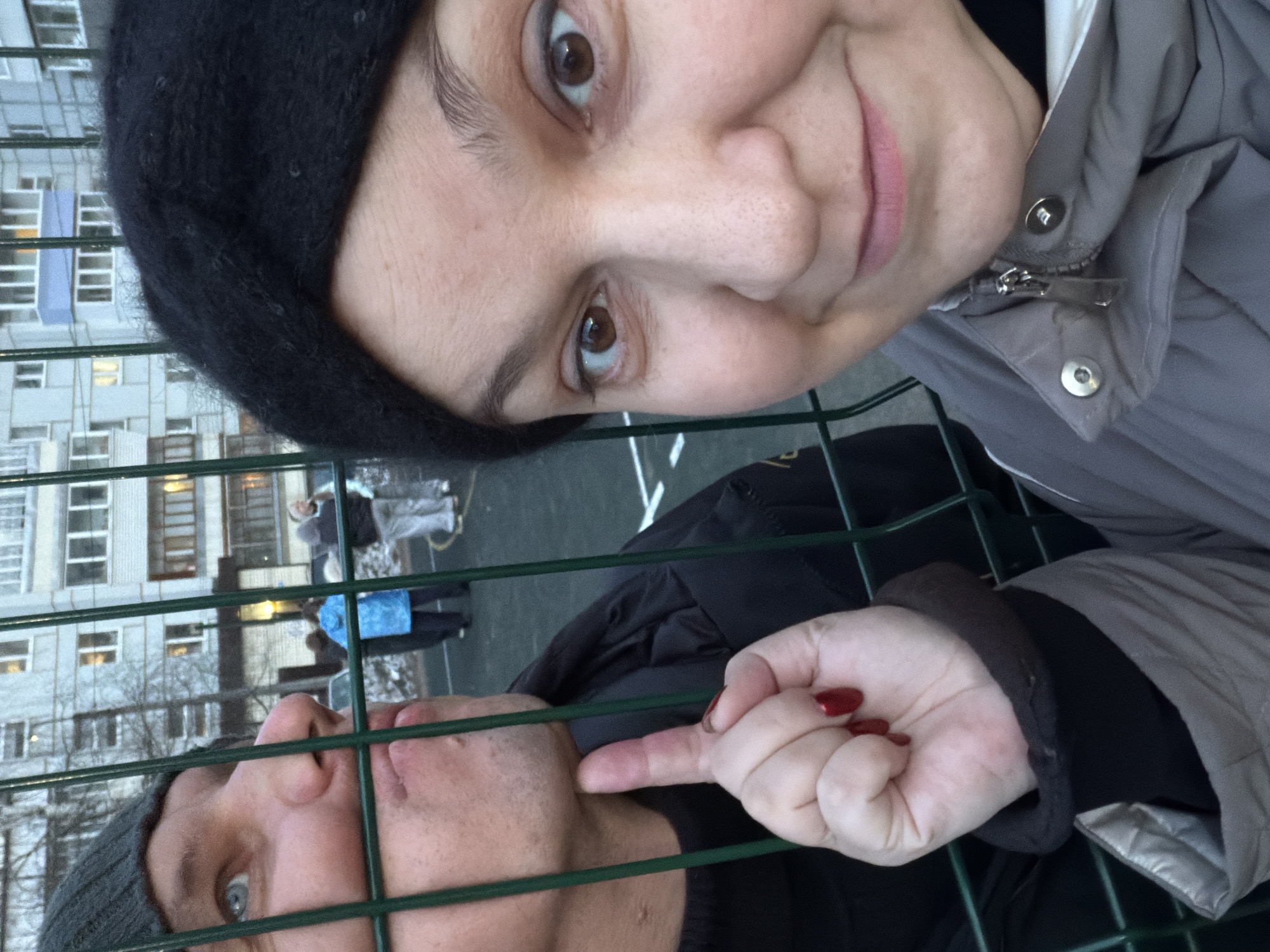 This portrait-oriented image, rotated 90 degrees clockwise, captures two individuals through a green wire fence, set against a backdrop of urban residential buildings.
In the upper section of the rotated frame, a woman is visible from the chest up, looking directly at the camera with a slight, friendly smile. She has brown eyes, dark eyebrows, and light skin, and is wearing a black knitted beret or beanie. Her attire includes a grey quilted jacket with a visible zipper and snap fasteners.
Below her, occupying the lower part of the frame, a man's face and upper torso are seen. He also looks towards the camera, wearing a dark grey or black knitted beanie and a dark jacket. He has blue-grey eyes and light skin with visible stubble. An interaction is taking place as the woman's hand, adorned with red nail polish, gently touches his cheek with her index finger, suggesting a playful or affectionate gesture.
A prominent green wire fence runs horizontally and vertically across the entire image, creating a grid pattern that frames the subjects. In the background, behind the fence, are several multi-story apartment buildings, characteristic of a residential urban environment. Some windows in the buildings show interior lights illuminated. A paved area is visible, and faint figures of other people can be seen in the distance, one wearing a bright blue coat.
The scene appears to be set during an overcast day, possibly late afternoon or early evening, given the ambient lighting and the illuminated windows. The outerwear worn by both individuals suggests cooler weather. The overall setting is consistent with a residential area in Chaikovsky, Russia. There is no visible text in the image.