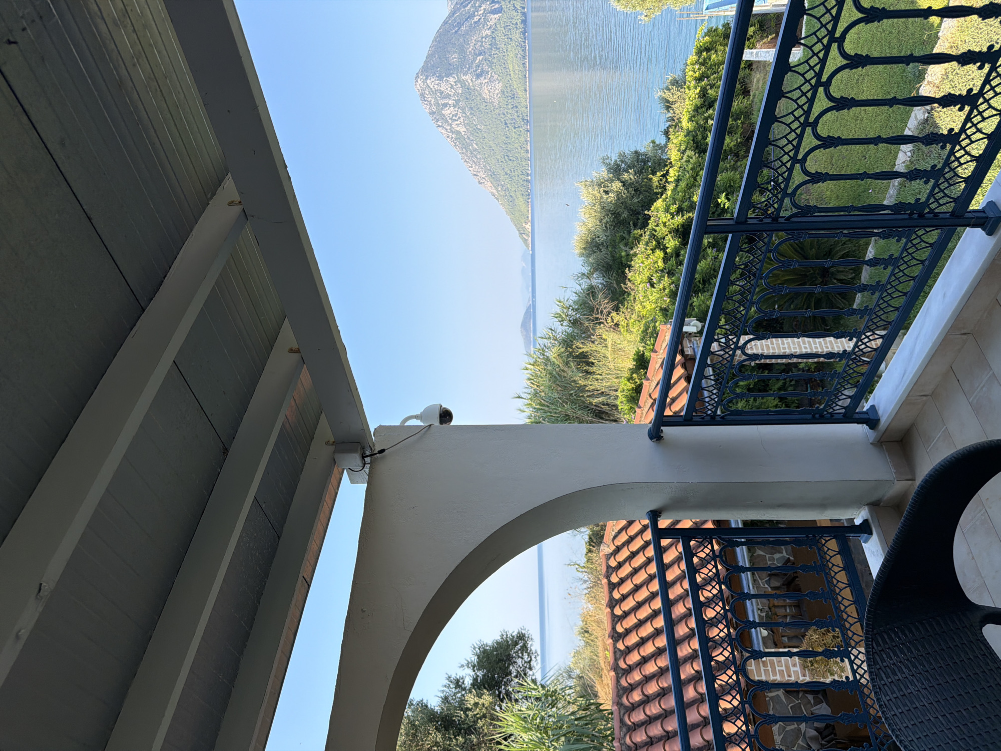 The image shows a balcony overlooking a body of water and a mountainous island. The balcony has a white railing with an ornate black metal pattern. A black wicker chair is visible on the balcony. Beyond the balcony, red-tiled roofs of buildings are visible, surrounded by lush green vegetation. The water is calm and reflects the clear blue sky. In the distance, a large, green, mountainous island rises from the sea. The scene appears to be in Municipal Unit of Alyzia, Greece. There are no people or animals visible in the image. The lighting suggests it is daytime.