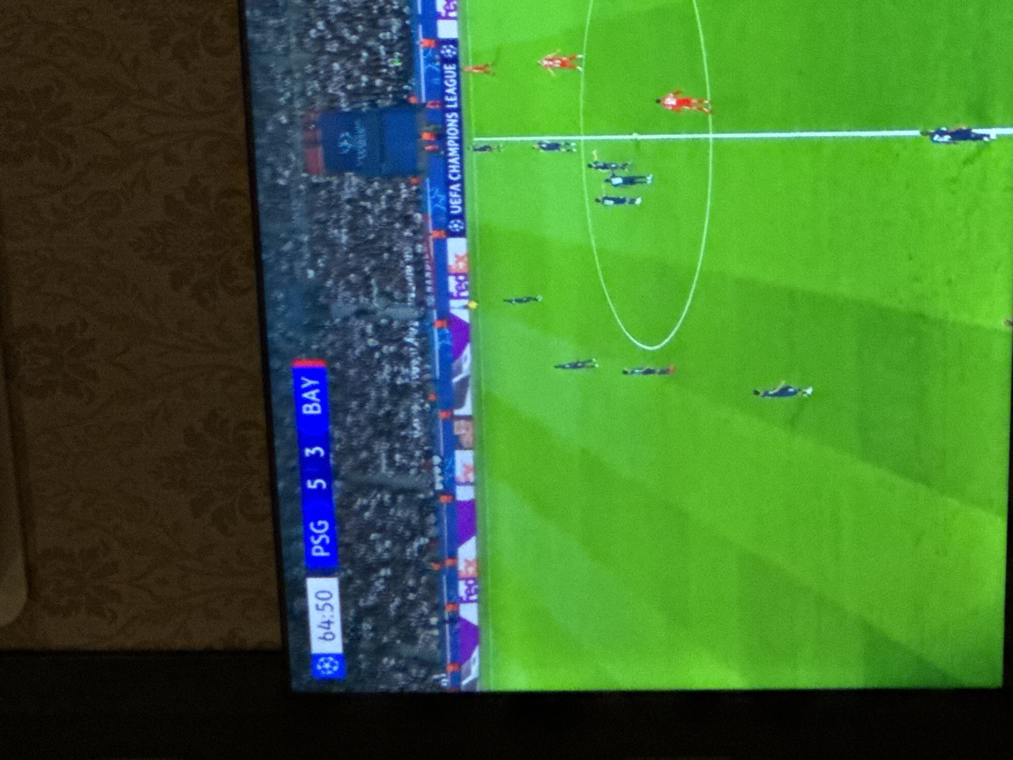 This image captures a television screen displaying a live football (soccer) match, viewed at an angle that rotates the screen clockwise. The screen's main subject is a vibrant green football pitch, marked with white lines and a central circle, indicating a standard playing field. Several players are visible on the pitch, appearing small due to the camera's perspective, with some wearing red kits and others dark blue or black kits.
Along the left side of the screen (which would be the top edge if viewed straight on), a crowded stadium stands is visible, though the spectators are blurred. A prominent scoreboard overlay is displayed vertically on the left side of the screen. It shows the match time as "64:50," indicating 64 minutes and 50 seconds into the game. Below the time, the scores are displayed: "PSG 5" and "BAY 3," meaning Paris Saint-Germain is leading Bayern Munich by five goals to three. A small UEFA Champions League logo is visible near the time and also along the advertising boards bordering the pitch.
Other visible text includes "UEFA CHAMPIONS LEAGUE" and the "FedEx" logo on the advertising hoardings. The television screen is set against a background of a wall covered in a textured, patterned wallpaper in shades of brown and beige, suggesting an indoor setting. The image captures an intense moment of a high-scoring UEFA Champions League match.
