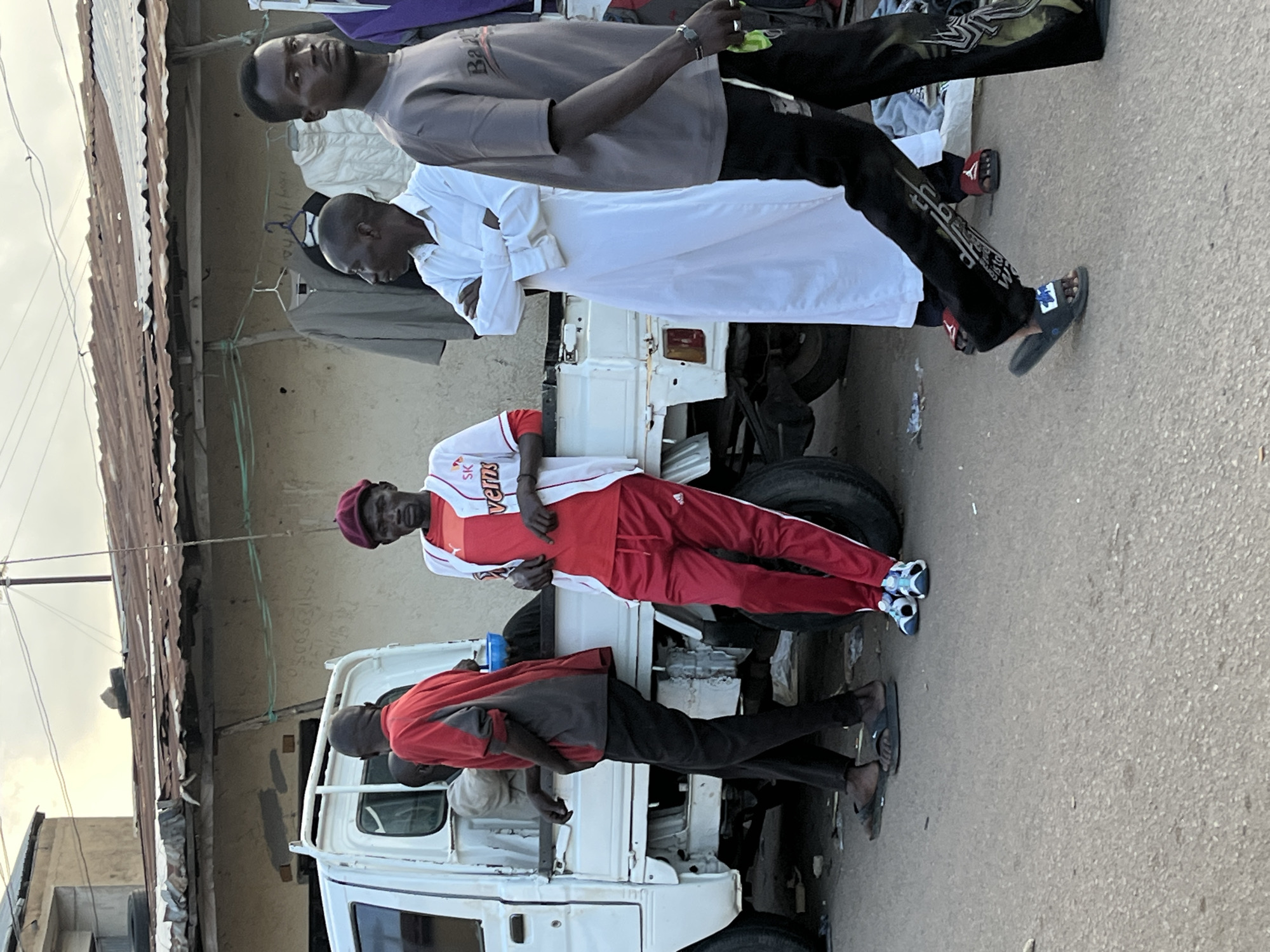 In Jos, Nigeria, a group of men stand near a white pickup truck. A man in a red shirt and black pants leans against the truck. Next to him, a man in a white baseball jersey with red accents and red pants stands facing forward. Further right, a man in a white robe with his arms crossed stands in profile. To the far right, a man in a grey t-shirt and black pants walks away from the camera. Behind the men, a market stall displays various clothing items. The scene is set on a paved street with a cloudy sky overhead.