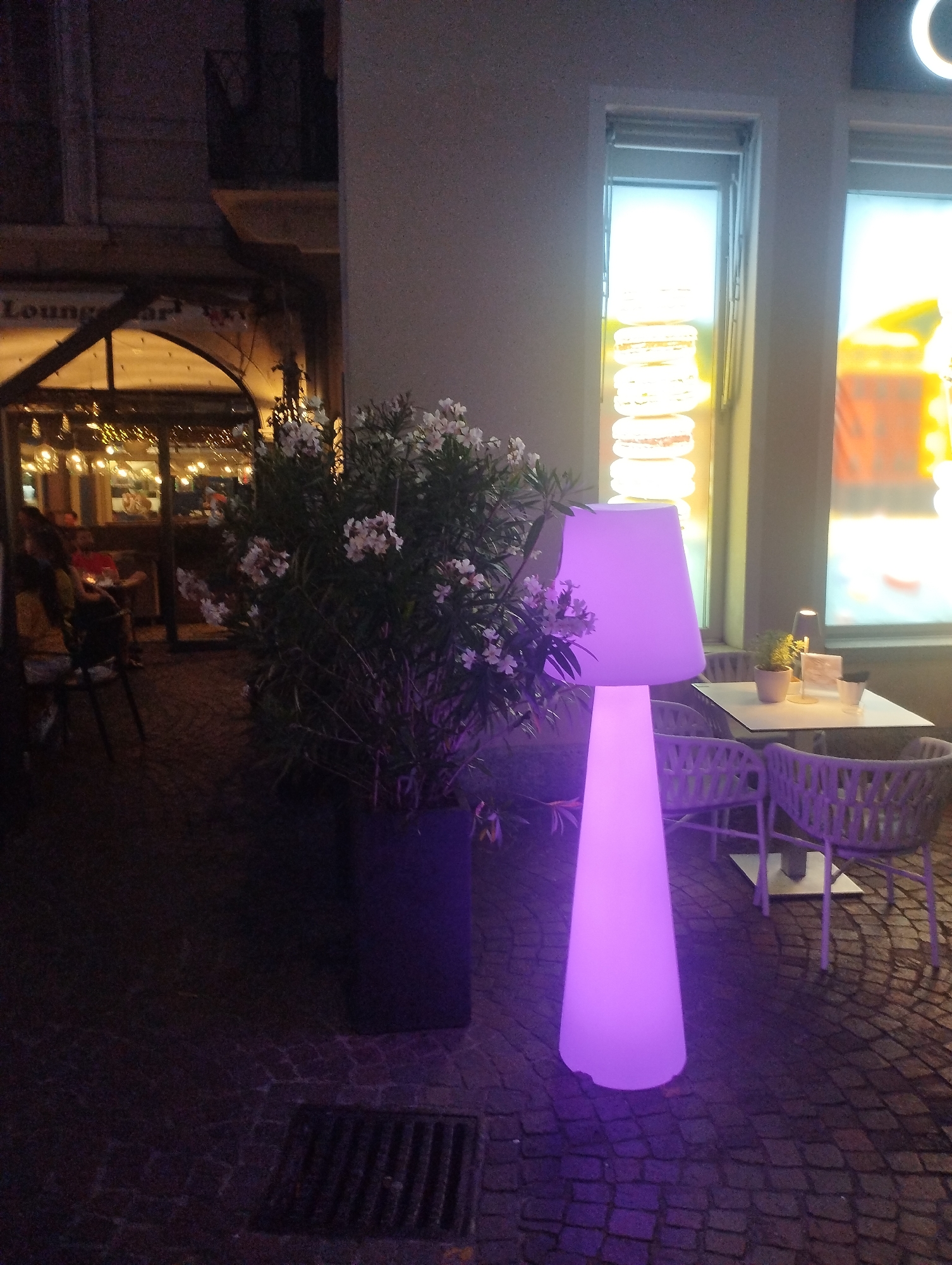 This image captures an evening scene on a cobbled street in Lugano, Switzerland, likely outside a cafe or restaurant. A large, cone-shaped lamp illuminates the area with a vibrant purple glow, casting shadows on the ground. To the left, an arched entrance leads into a building, where lights and figures are visible inside. A lush green bush with small white flowers is planted in a dark planter, also bathed in the lamp's purple light. In the foreground, outdoor seating areas with tables and chairs, designed in a modern, woven style, are arranged. One table is set with small potted plants and what appear to be glasses. A grate is visible in the pavement in the lower left corner.