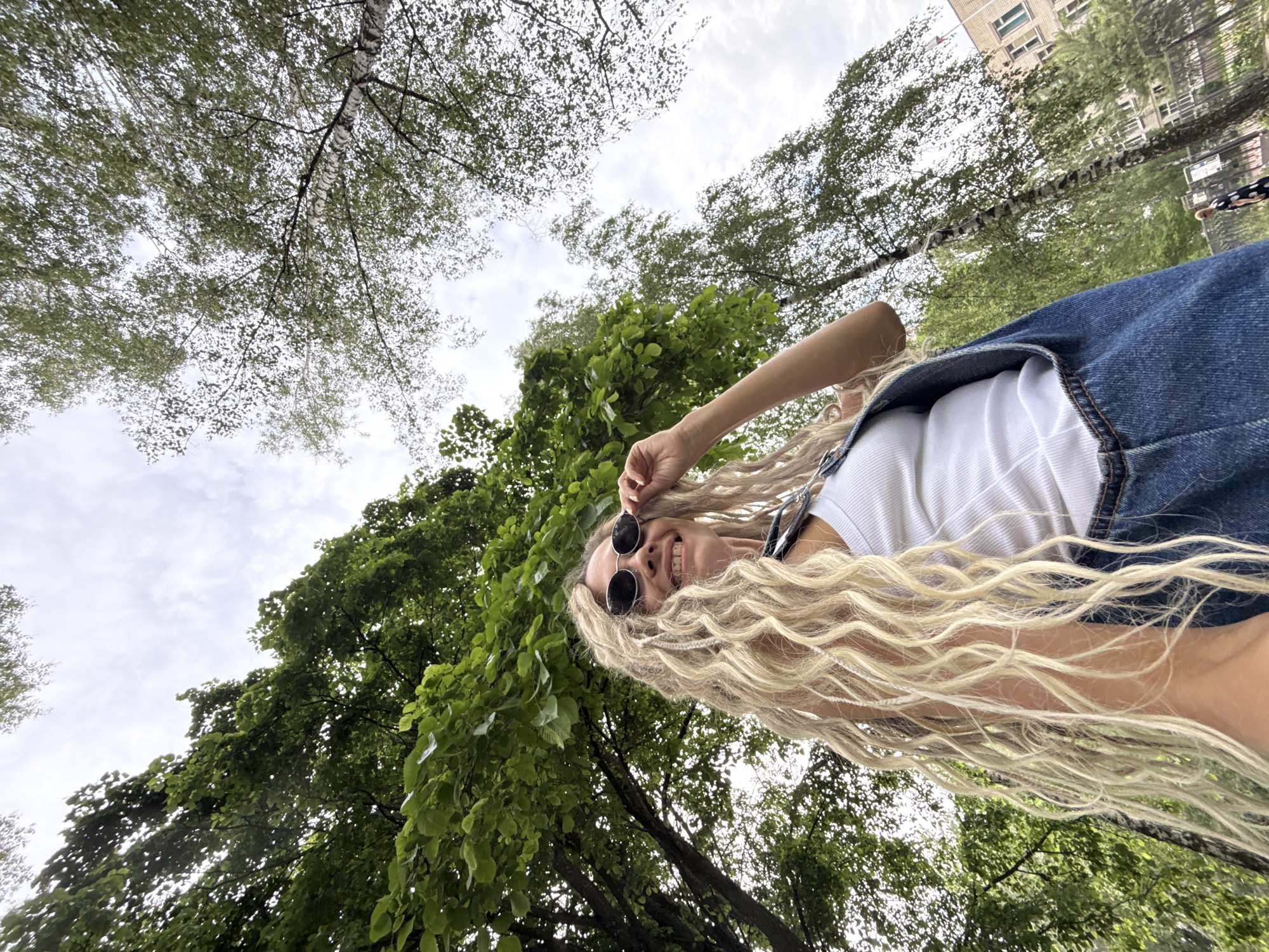 A young woman with long blonde dreadlocks and sunglasses is standing outdoors in what appears to be a park or green space, possibly in Rusanovo, Russia. She is wearing a white tank top and denim overalls. The camera angle is low, looking up at her against a backdrop of lush green trees and a cloudy sky. In the background, a building is visible, suggesting an urban or semi-urban setting. There is a hint of another person in the far background, walking.