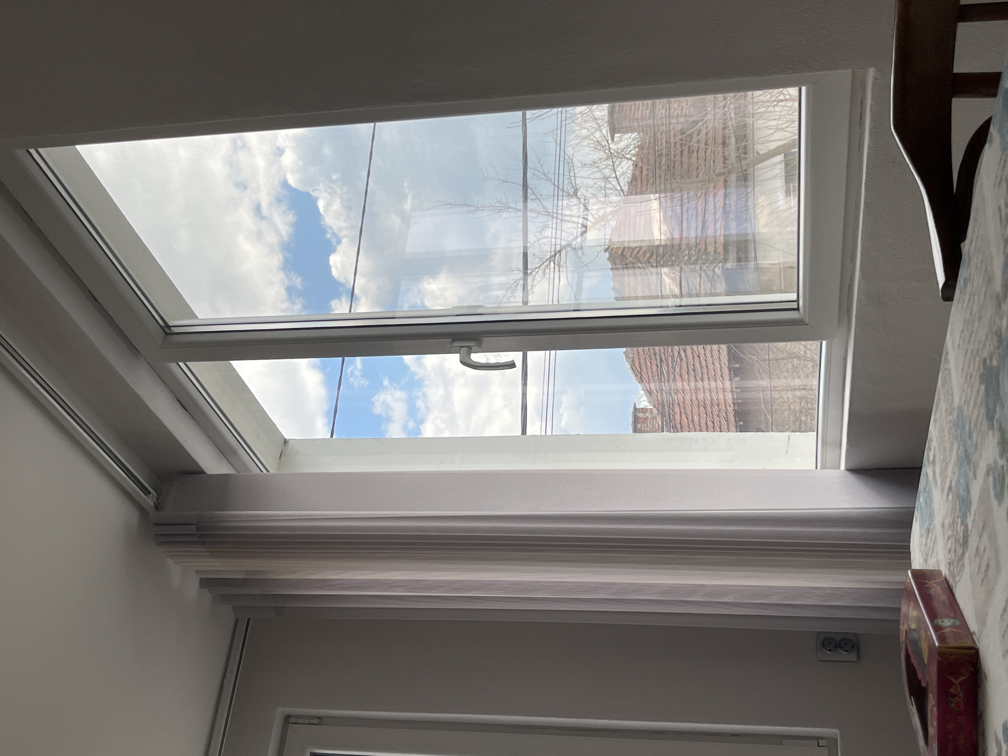The image shows an interior view of a room, likely in a house, featuring a rectangular skylight. The skylight frame is white, and it appears to be open slightly, revealing a view of a partly cloudy blue sky and a reflection of buildings with red-tiled roofs, possibly of residential structures, and bare tree branches. The room’s walls are painted a light color. Below the skylight, there is a closed, layered blind or shade of a neutral color. A portion of a chair back is visible on the right side of the image, with a patterned fabric nearby. A small, rectangular object is situated to the right of the bottom of the shade, close to the wall. No people are visible in the scene. The presence of red-tiled roofs suggests a location with Mediterranean architectural influences, consistent with Bulgaria. No specific details in the image confirm or deny the location being in the city of Karavelovo, Bulgaria.