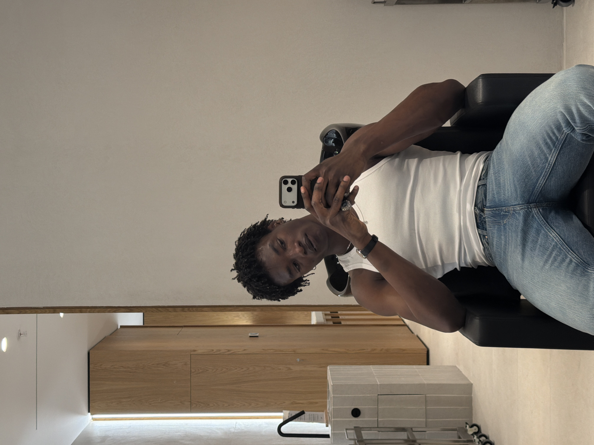 A man is taking a selfie while sitting in a black chair in a room with modern design. The room has light-colored walls, a wooden cabinet, and recessed lighting. The man is wearing a white tank top and light blue jeans. He has short, dark hair. He is holding a smartphone in his left hand, with his fingers wrapped around the phone and his thumb extended. He is looking at the camera with a neutral expression. The background of the image appears to be a sleek and minimalist interior. The location is in the city of Fibracolor, Spain.