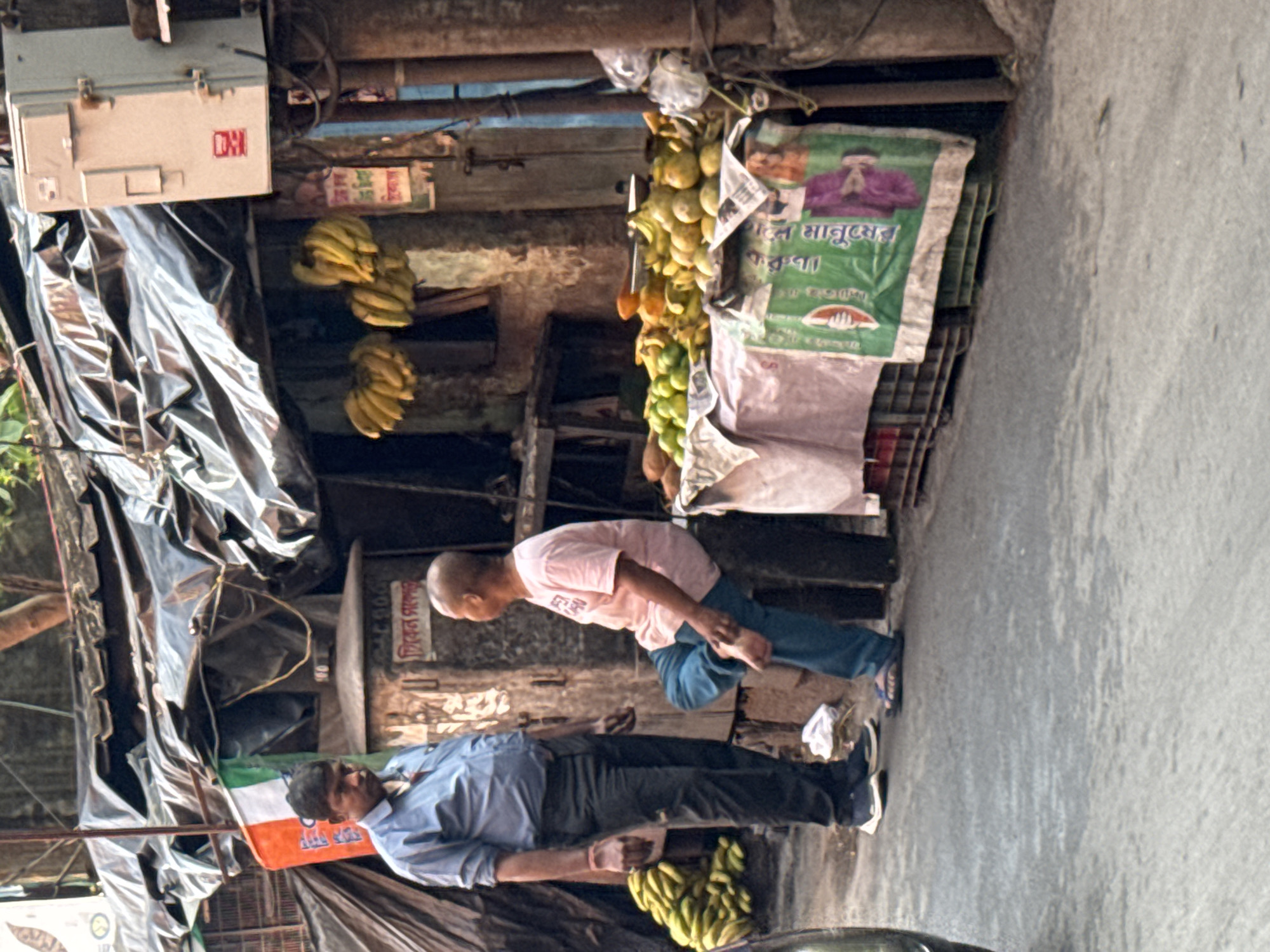 The image shows an outdoor street scene in Kolkata, India. Two men are the main subjects. One man is wearing a light blue shirt and dark pants and is standing near a small pile of bananas on the ground. The second man, who is bald and wearing a light pink shirt and blue jeans, is bending over. A fruit stand with hanging bunches of bananas and mangoes is visible in the background. There is an Indian flag draped to the side. The environment is urban, with a building façade and black tarpaulin sheets covering structures.