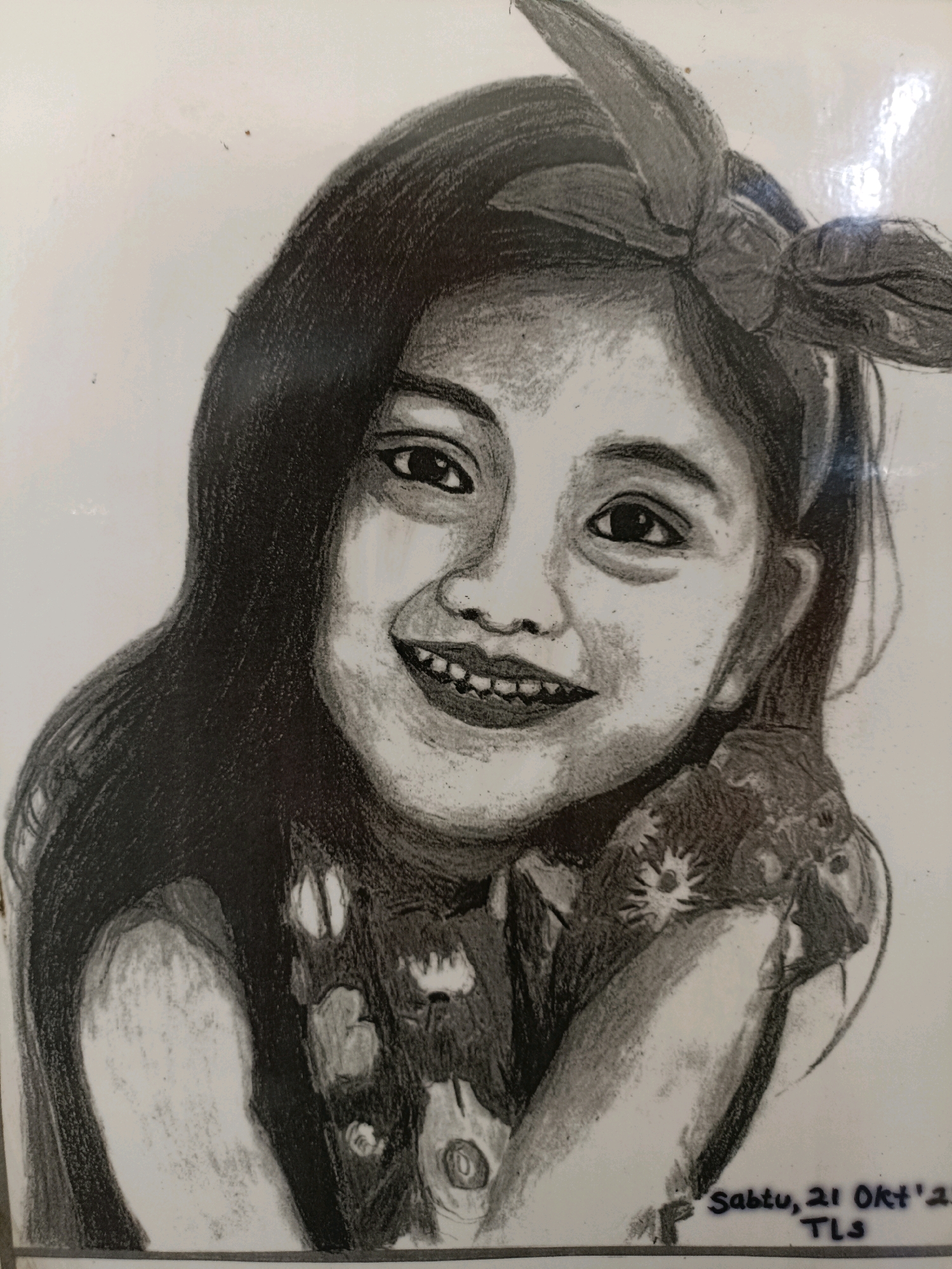 A charcoal sketch depicts a young girl with a cheerful expression, her head tilted slightly and her hands resting near her face. She has dark, long hair styled with a bow on top and wears a patterned top. The sketch is signed and dated "Sabtu, 21 Okt '21 TLS" in the lower right corner. The background is white and appears to be a wall. There are some small dark specks on the white surface, possibly dust or minor imperfections. The image is taken indoors, likely in a home environment in Surabaya, Indonesia, given the Indonesian date inscription.