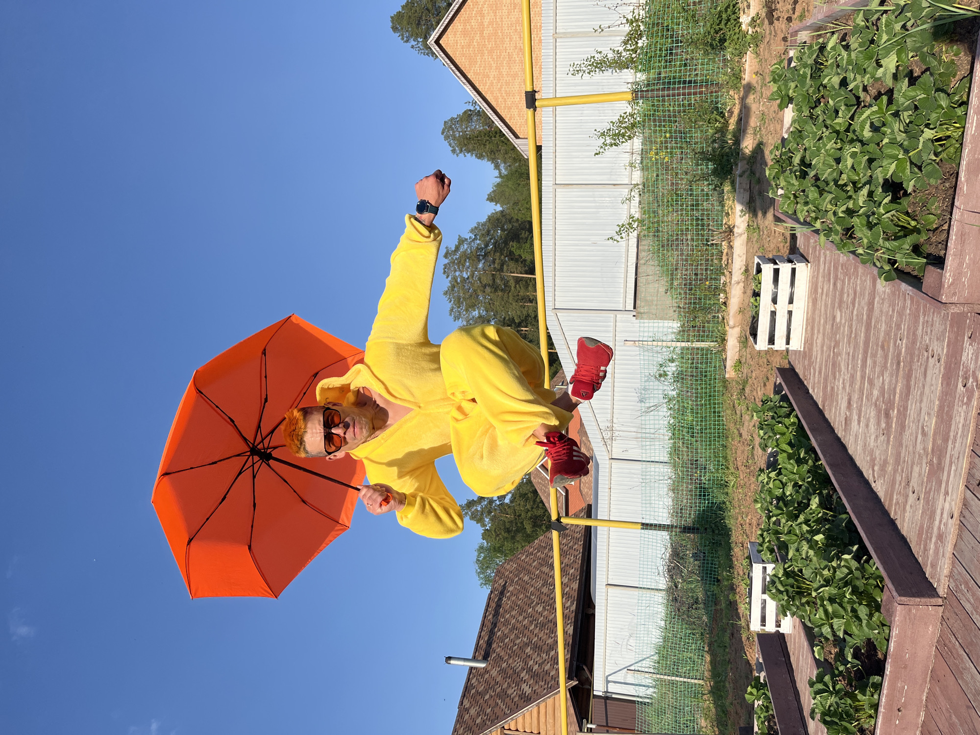 A man is depicted in mid-air, holding an orange umbrella. He is wearing a yellow hooded jumpsuit and red sneakers. The backdrop includes a clear blue sky, portions of houses with varying roof colors (brown and brick), a white fence with a wire mesh top, greenery, and raised garden beds. The location appears to be in Chaikovsky, Russia, based on contextual information.