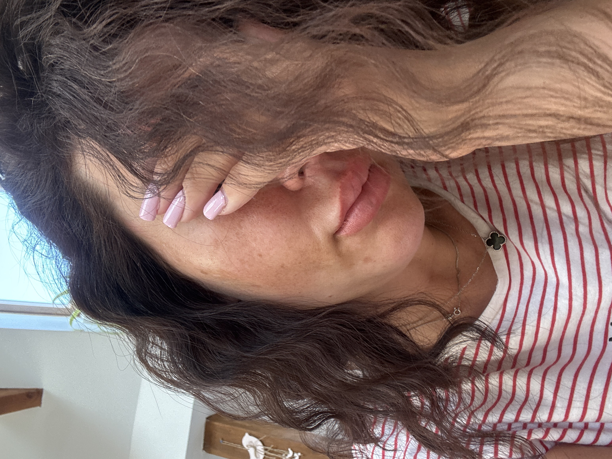 The image shows a close-up, medium shot of a woman's face, partially obscured by her hand and dark, wavy hair. She is wearing a red and white striped t-shirt and a delicate silver necklace with a clover-shaped pendant. Her skin has visible freckles and appears slightly flushed on her cheek. Her fingernails are painted a light pink. The lighting suggests an indoor setting, possibly near a window, indicated by a bright light source and a hint of greenery in the background. There is no identifiable information to determine the location of the image within the city of Katowice, Poland.