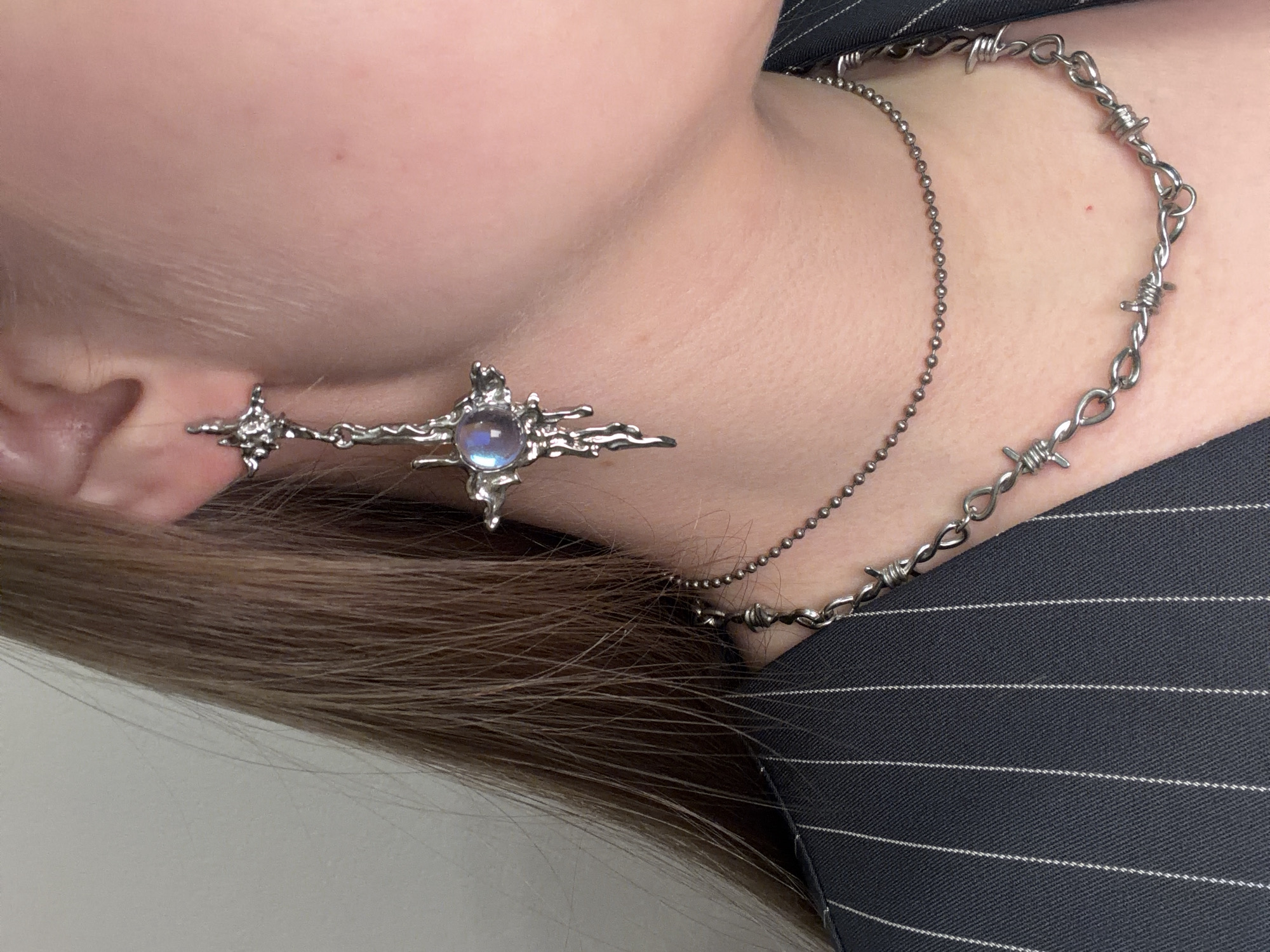 A close-up, angled shot captures the neck, ear, and shoulder of a person, focusing on their jewelry and fashion. Long, brown hair is visible, falling over the shoulder.
The person wears a striking metallic earring on their left ear, featuring an irregular, organic star-like or cross-like design. This earring is prominently set with a central, iridescent stone that displays a luminous, opalescent sheen, resembling a moonstone. A smaller, simpler metallic star accent is attached further up the ear.
Two distinct silver-toned necklaces adorn the person's neck. One is a delicate ball chain, while the other is a thicker, more assertive chain designed in the style of barbed wire. The person is dressed in a dark-colored garment, possibly a jacket or top, characterized by fine white pinstripes visible on the shoulder area.
The background is a plain, light-colored surface, suggesting an indoor setting. There is no discernible activity taking place, and the image appears to be a stylistic shot highlighting personal accessories. No text is visible in the frame.