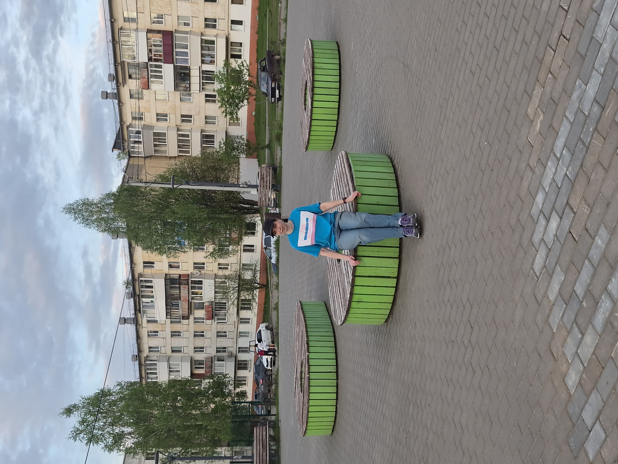 A medium, vertical shot shows a person resting on a green and wood bench located in Chaikovsky, Russia.
The person, presumed female, is dressed in blue and jeans with a cap, lying across the bench, facing the viewer. The bench is circular, made of green painted vertical planks with a wood top, and there are two identical benches on either side of the center bench.
The setting appears to be a paved public area, possibly a park or common area. Buildings are visible in the background. Several cars are parked. A low fence can be seen in front of the building. The weather is overcast.