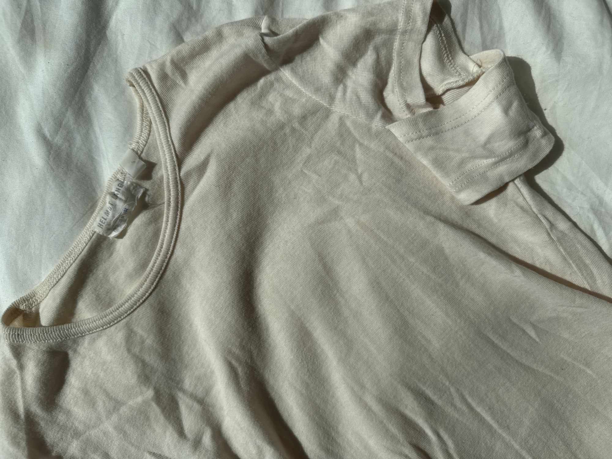 The image displays a cream-colored short-sleeved garment, likely a t-shirt or top, laid flat on a light-colored, possibly white, wrinkled fabric surface. The garment occupies the majority of the frame, positioned diagonally with its upper section towards the top left and its main body extending towards the bottom right.
Strong, natural light illuminates the scene, casting distinct shadows across both the garment and the background fabric, suggesting it is daytime, possibly morning or late afternoon. The garment is made of a soft, knit material, with visible details including a ribbed crew neck collar and a cuffed sleeve visible on the right side.
A white fabric label is affixed to the inner left shoulder/neck area of the garment. The text "HELMUT LANG" is clearly visible and legible on this label. Below this, partial text is visible that likely indicates the country of origin.
The background fabric, with its folds and creases, provides a subtle textural contrast to the garment. The overall color scheme is muted, featuring various shades of cream, off-white, and the light browns and grays created by the shadows. There are no people visible, and no activity is taking place; the scene is static, highlighting the textile and its branding under natural light. The setting appears to be an indoor environment, such as a room or studio in London, United Kingdom.