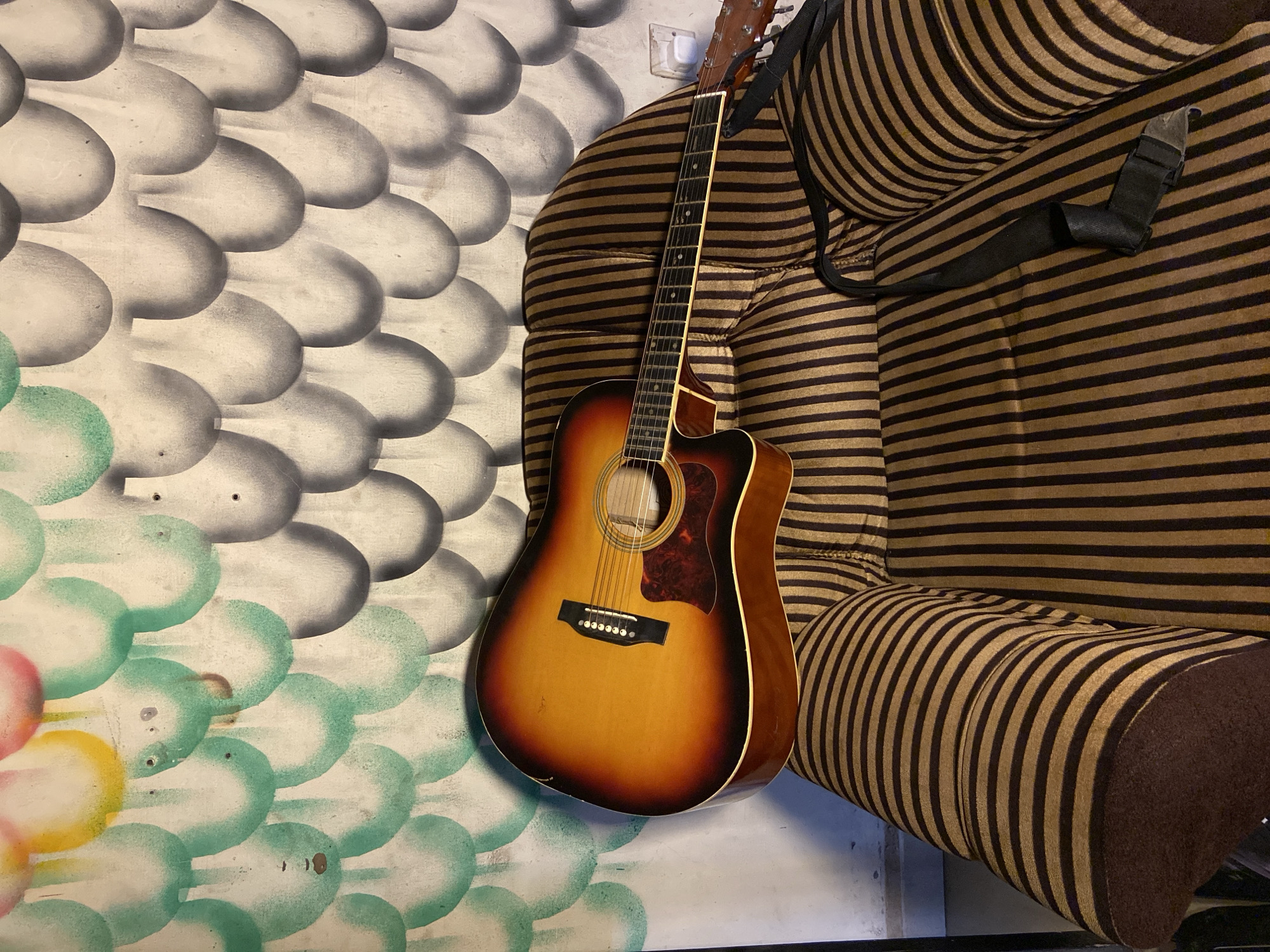 An acoustic guitar with a sunburst finish leans against a patterned wall and a striped couch in an indoor setting in Mararaba, Nigeria. The guitar, prominently featured, displays a warm brown-to-orange gradient on its body, a dark pickguard, and a light fretboard adorned with dot inlays. A black fabric strap is draped over the adjacent furniture.
The background wall is decorated with a distinctive, hand-painted pattern resembling overlapping scales or bubbles. The upper portion of this design is rendered in grayscale, while the lower section transitions into vibrant hues of teal, green, yellow, and hints of pink, suggesting a decorative or artistic wall treatment. The painted surface shows some minor wear and imperfections. A white electrical outlet is visible on the wall behind the guitar's headstock.
To the right, a plush couch or armchair is upholstered with alternating horizontal stripes of dark brown and a lighter, sandy brown or gold color. The guitar is positioned between the textured, artistic wall and the boldly striped furniture. No activity or individuals are present in the image. The lighting is even, typical of an indoor environment. There is no visible text.