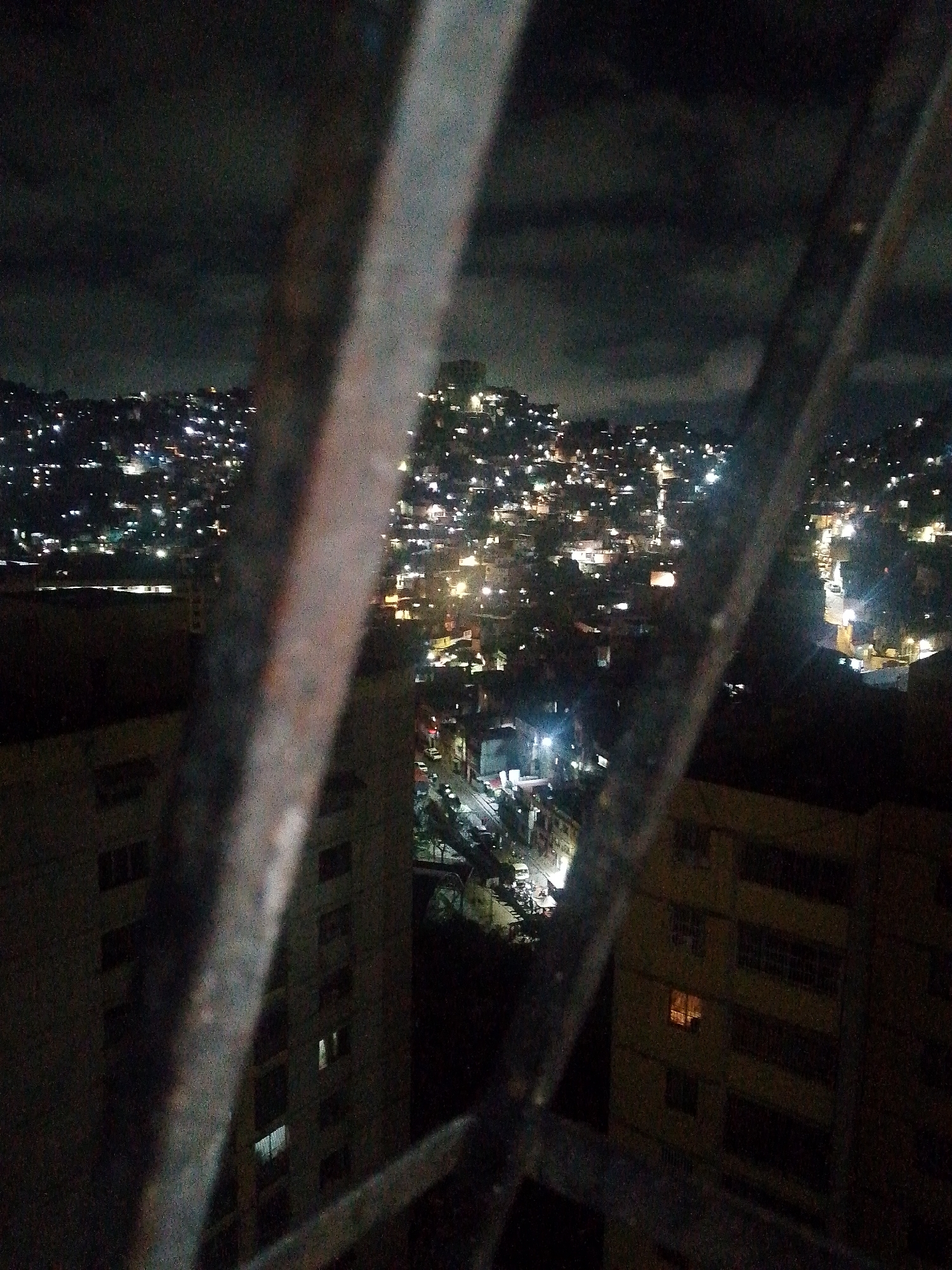 The image is a nighttime view of Caracas, Venezuela, from behind a barred window. The city sprawls across a hilly landscape, illuminated by numerous lights. Two buildings are visible in the foreground, one on either side of the window. The bars of the window are thick and rusty, obscuring the view. The overall atmosphere is dark and gritty. The specific location within Caracas cannot be determined without additional information.