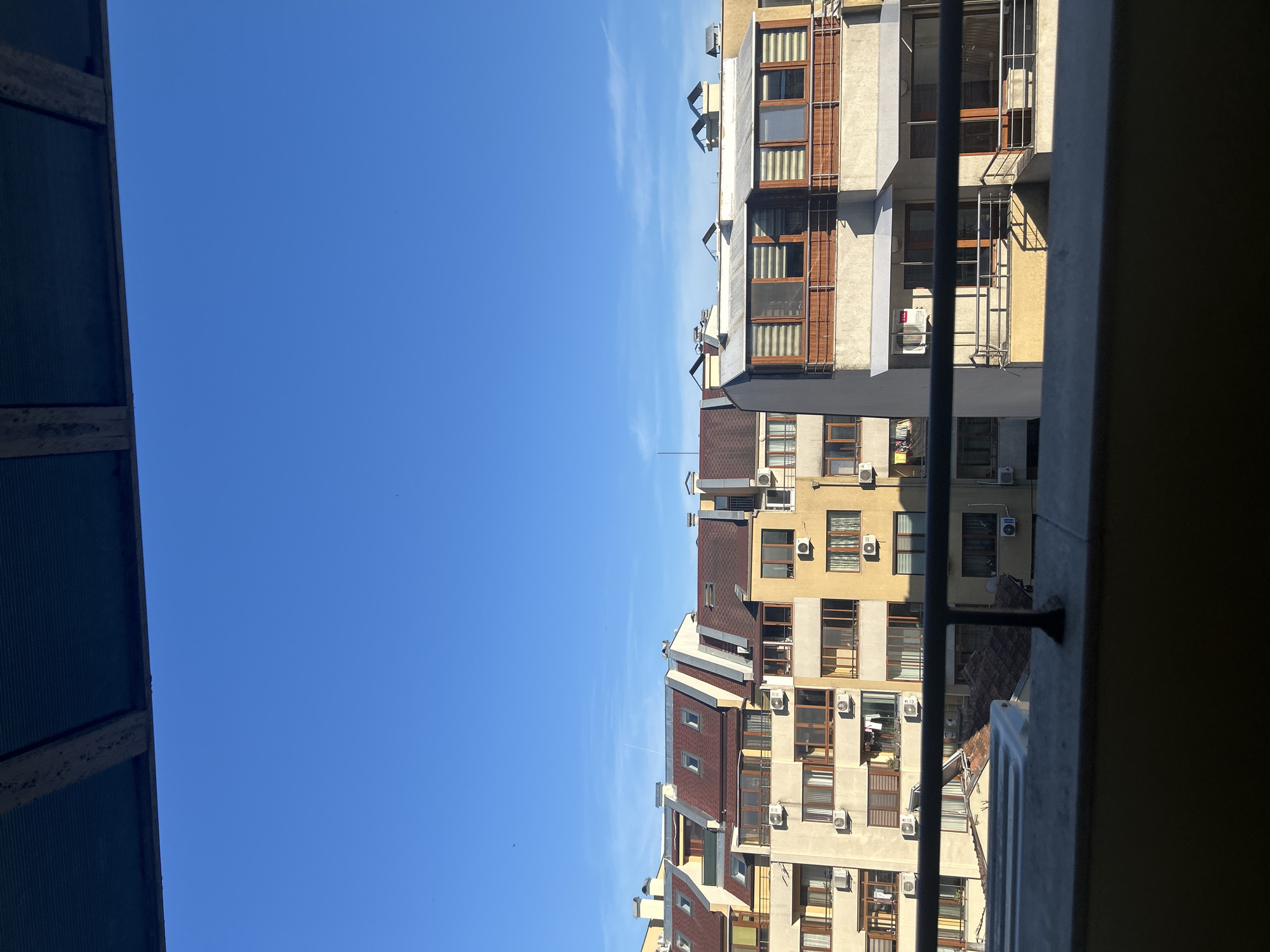 The image depicts a view of apartment buildings in Varna, Bulgaria, under a clear blue sky. The buildings are multi-story structures with varied architectural features, including red-tiled roofs, balconies with railings, and numerous windows and air conditioning units. Sunlight casts shadows on the facades, indicating it is daytime. The foreground shows a railing and part of a concrete structure, suggesting the photo was taken from a balcony or window. The scene is urban and residential, with no people visible.
