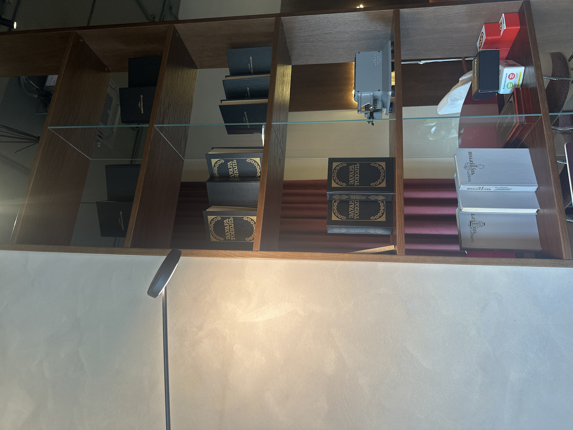 Here is a detailed description of the image:
The image is a shot of a shelf with a variety of objects. The shelf is made of wood and has a glass covering. The objects on the shelf include books (some with titles like "EDUARD TOPOL", "GILBERT K. CHESTERTON" and "Файхтвангер" (Feuchtwanger)), and board games. Also visible are what appears to be an antique projector and a lamp in the lower left part of the image.
It's difficult to determine the precise location in Stavropol, Russia with just the information provided, as the specific location would require recognizable landmarks. The scene suggests an indoor setting, potentially a study, library, or a retail space.