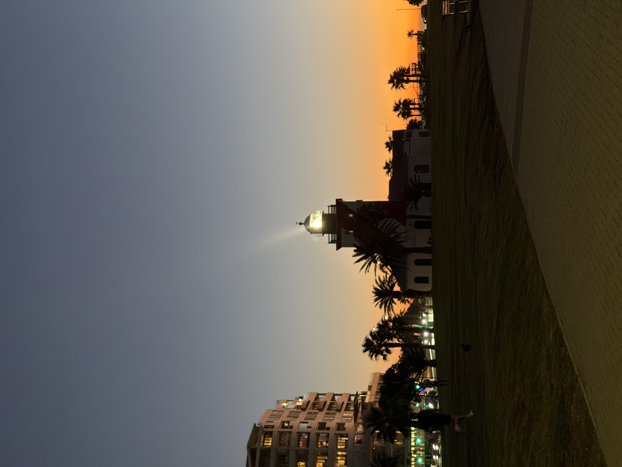 Here is a description of the image:
The image shows a lighthouse at dusk, probably the Mouille Point Lighthouse in Cape Town, South Africa. The lighthouse is lit, shining a beam into the sky. The sky above is a gradient from dark blue to orange. There are other buildings and palm trees silhouetted against the sky. A walkway or pavement borders the bottom right of the image. People are seen near the bottom center of the image.