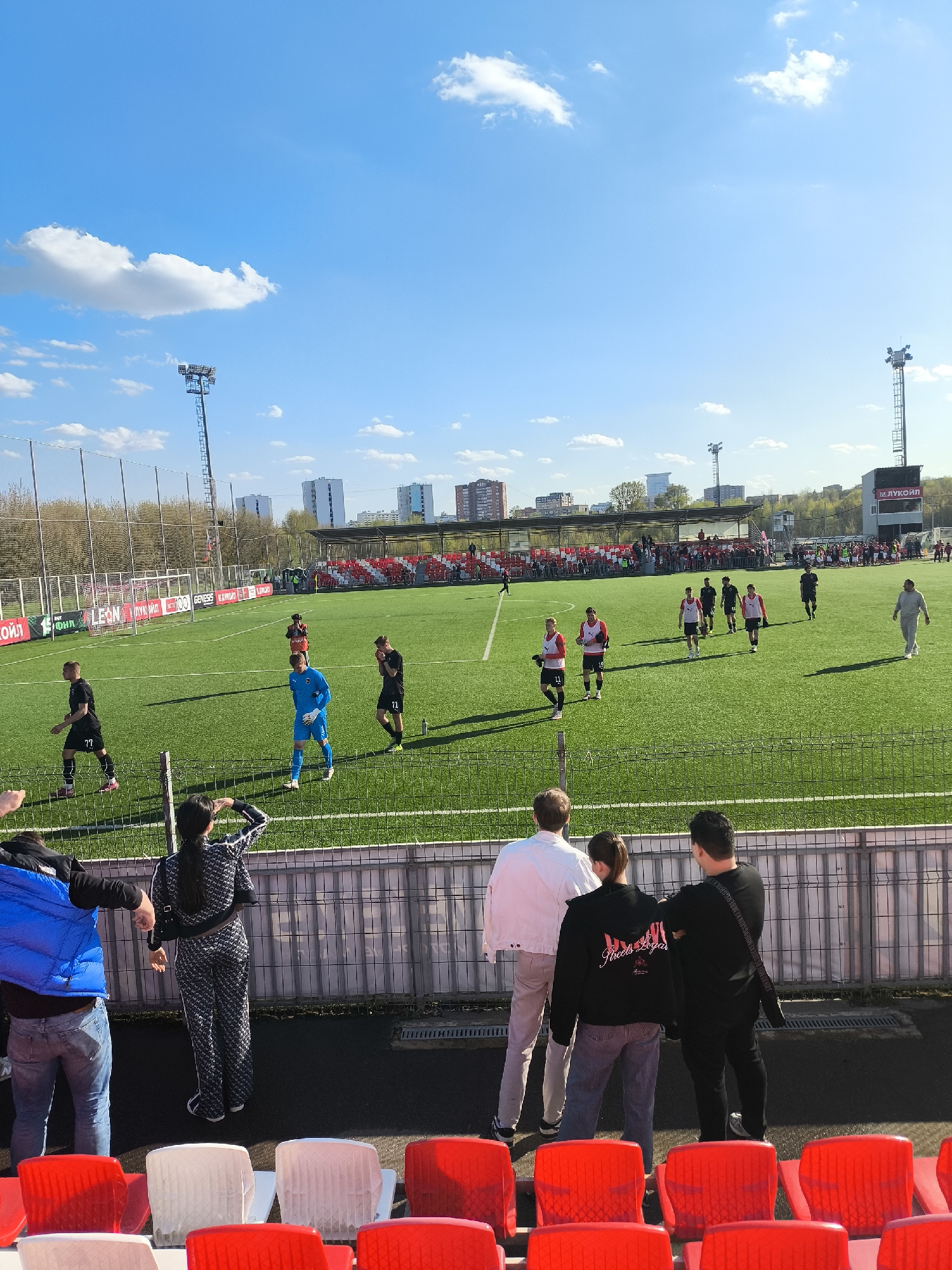 The image depicts a soccer game in progress, likely in Moscow, Russia, based on the "LUKOIL" sign visible on the stadium scoreboard. The stadium is filled with spectators, particularly in the red and white colored stands in the background.
The game is being played on a green field with white lines marking the boundaries. Several players are visible, wearing different colored jerseys. There appears to be a team in red and white jerseys, and another in black. One player is wearing a bright blue goalkeeper uniform.
In the foreground, several spectators are standing behind a barrier, watching the game. A woman is wearing a black and white patterned outfit. Another person is wearing a white jacket, and another a black hoodie with graphic art. Red and white plastic chairs are visible in the foreground, suggesting a seating area for spectators.
Tall light towers can be seen above the stadium, with a partially cloudy blue sky in the background. Residential buildings can be seen in the distance beyond the stadium.