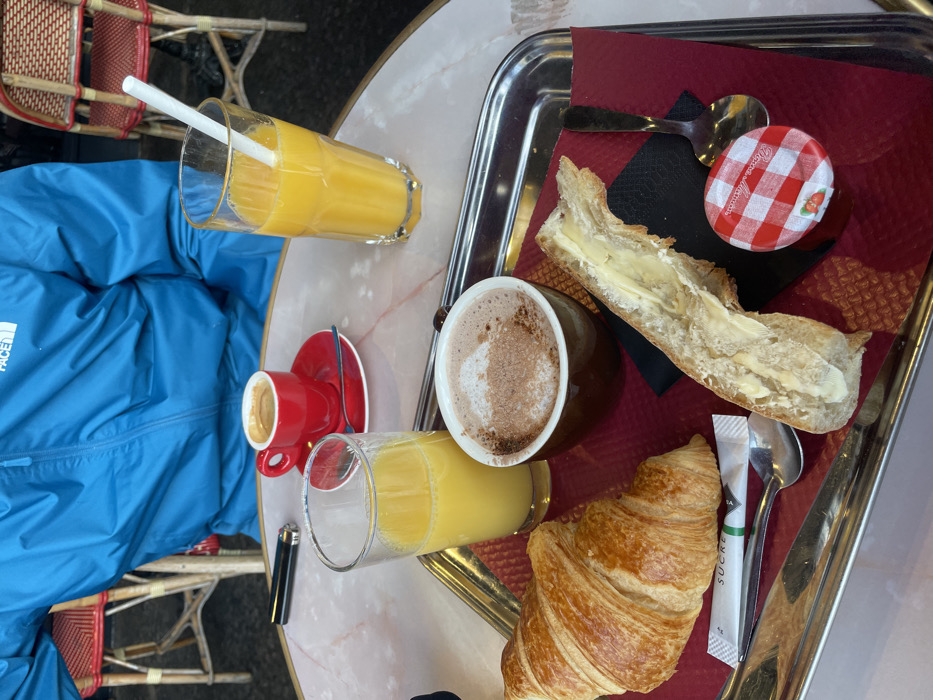 The image displays a breakfast or brunch scene at an outdoor café in Paris, France. A silver rectangular tray occupies the central foreground, placed on a light-colored, possibly marble, round café table. The tray holds a red textured placemat and an assortment of food and beverages. On the tray, there is a golden-brown croissant, a segment of baguette buttered and split lengthwise, a small brown ceramic mug containing hot chocolate dusted with cocoa powder, a small glass filled with orange juice, a small individual jar of strawberry jam with a red and white checkered lid, a silver spoon, a white sugar packet labeled "SUCRE," and a small black napkin.
Off the tray, on the table, a smaller glass of orange juice is positioned to the left. Behind the tray, a vibrant red espresso cup sits on a matching red saucer with a tiny spoon. A black lighter rests to the left of the espresso setup. To the right, a taller glass of orange juice contains a white straw.
A person, partially visible, is seated behind the table, wearing a bright blue jacket, identifiable by a partially obscured "The North Face" logo. The individual's face is not visible. In the blurred background, classic Parisian café chairs with light wooden frames and red woven cane seats are discernible, indicative of a typical street-side café setting.