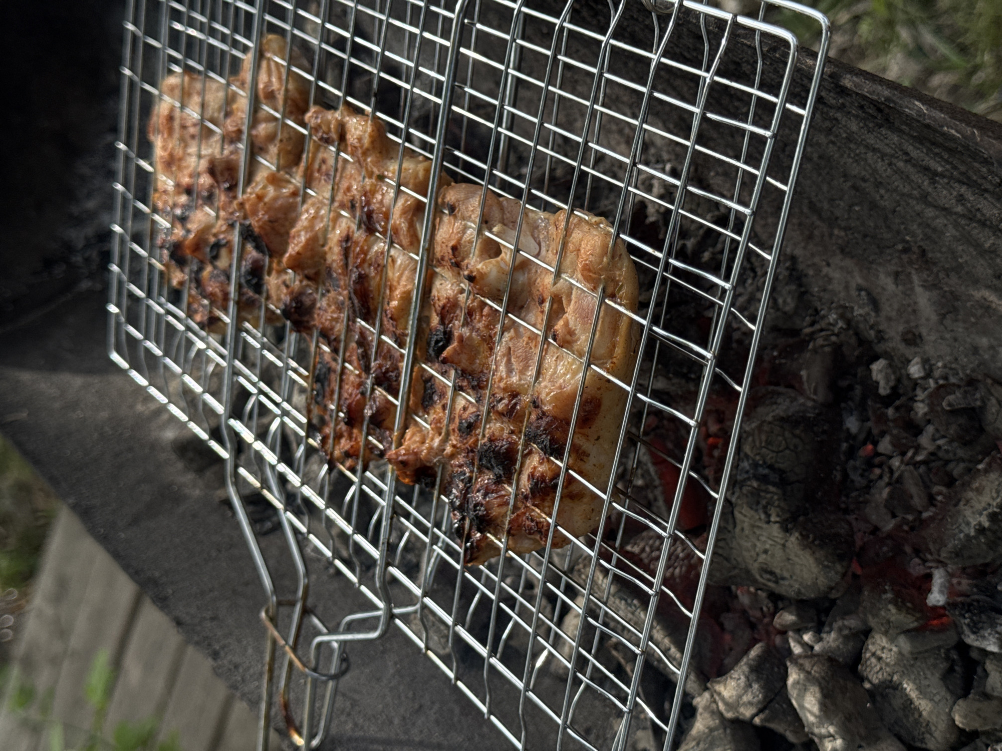 A close-up shows grilled meat, presumably chicken, cooking in a wire grilling basket over coals. The meat is browned and slightly charred. The grilling basket sits over a bed of gray and black coals, with some glowing red. The scene appears to be outdoors, with some indistinct vegetation visible.