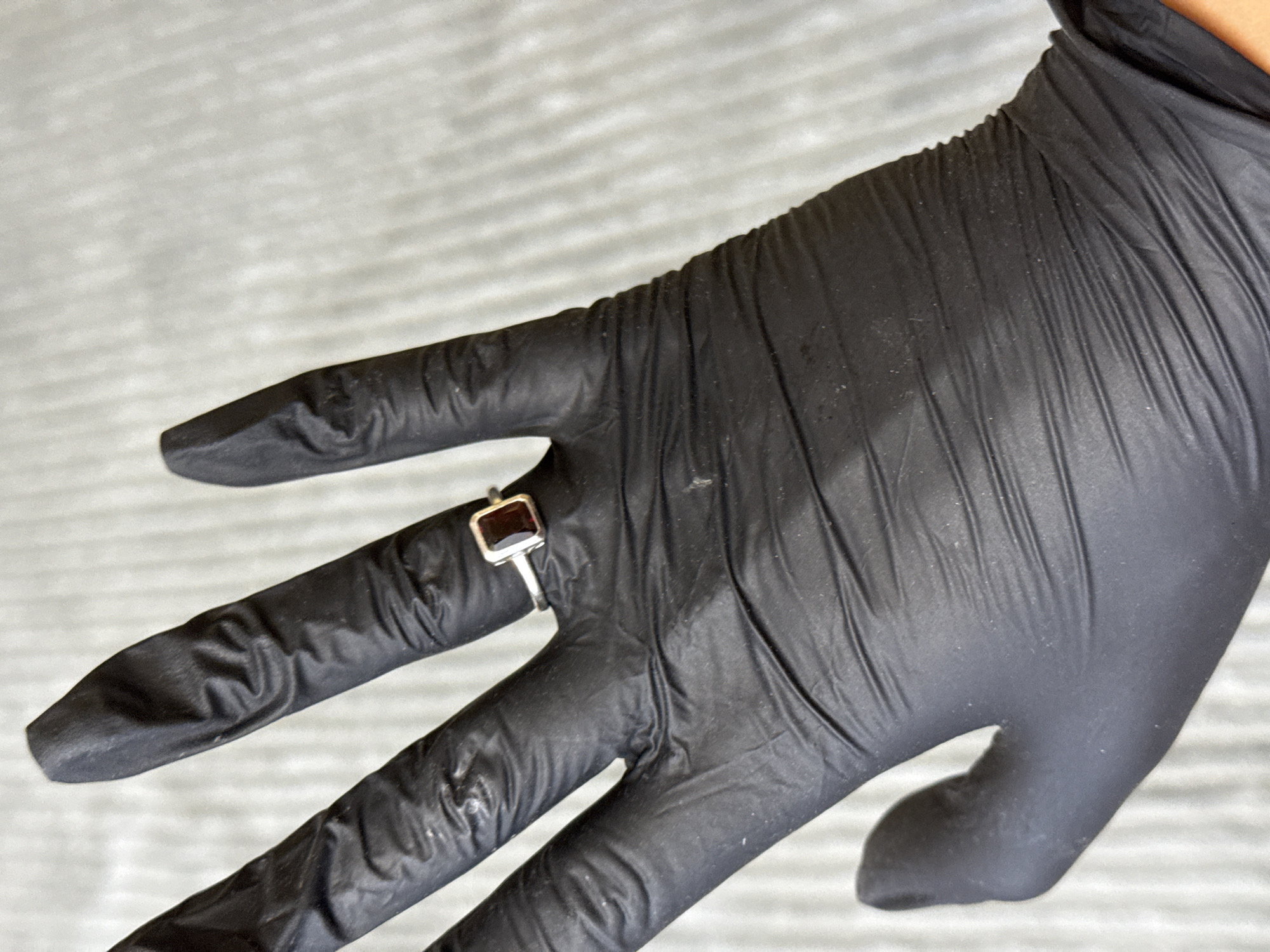 This is a close-up, eye-level shot of a gloved hand displaying a ring. The hand is covered in a black latex or nitrile glove, which is creased and shows some texture. The ring, worn on the index finger, is silver with a rectangular, dark red gemstone. The background is a light gray, textured surface, possibly fabric or paper, with horizontal striations. The lighting appears even, suggesting an indoor setting with artificial light. There is no visible text or discernible time of day or weather conditions. The focus is on the ring and the gloved hand, creating a still-life composition.