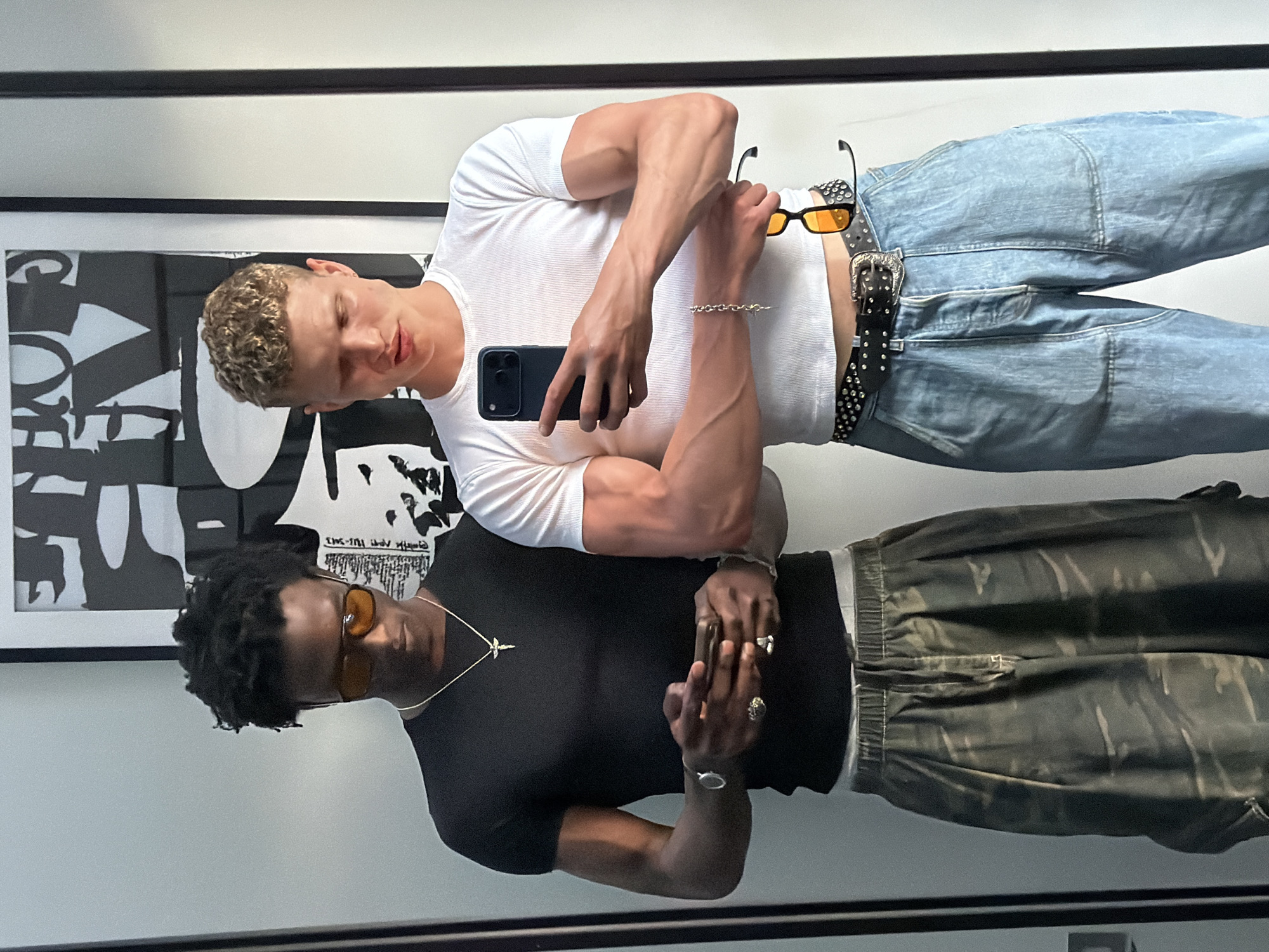 Two men are taking a mirror selfie. One man is light-skinned, wearing a white t-shirt, light-wash jeans, a studded belt, and is holding yellow-tinted sunglasses. The other man is dark-skinned, wearing a black t-shirt, camouflage pants, and a gold necklace with a cross pendant. Both men are wearing rings and bracelets. The background includes a black and white framed poster and a white wall. The location is assumed to be indoors, in London, United Kingdom.
