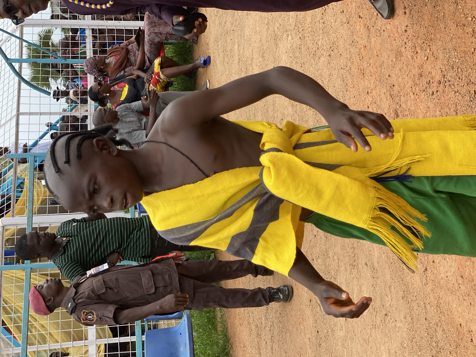 The image shows a young, dark-skinned boy with braided hair standing in an outdoor setting in the city of Maigizo, Nigeria. He is bare-chested and wearing a yellow and grey striped scarf-like garment around his torso and green pants. The ground is dirt or sand. Behind him are several people seated in a metal-framed enclosure, possibly a spectator area. A man in a brown uniform and red cap is standing to the boy's left, partially obscured. In the background, more people can be seen, some sitting and some wearing traditional African clothing.