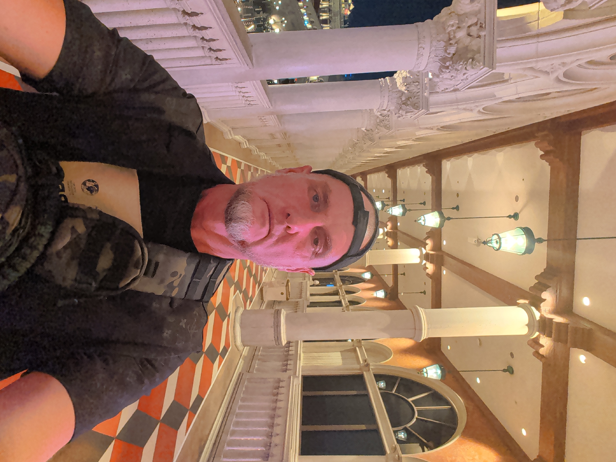 The media file shows a selfie of a man in what appears to be an indoor setting with architectural details reminiscent of old European or Venetian style. The man is wearing a black t-shirt with a camouflage vest over it and a black cap. He has a goatee and is looking towards the camera. The background features ornate columns and railings, possibly from a balcony or walkway. The floor has a distinctive diamond pattern with orange, grey, and white tiles. Hanging lamps with green glass shades are visible, suggesting an interior space with decorative lighting. The overall impression is that of a tourist taking a photo at a hotel or resort, possibly in the evening or at night given the interior lighting and the distant view which seems to be illuminated buildings against a dark sky. The man's expression is neutral. There is text on a light brown tag visible on the man's shirt, but it is not clearly legible. The location is identified as Paradise, United States of America.