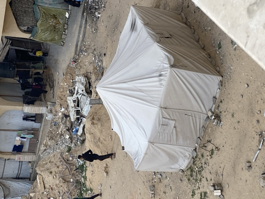 The image captures an outdoor scene, rotated approximately 90 degrees counter-clockwise, depicting a makeshift encampment or an area affected by disarray, viewed from a high vantage point looking downwards.
In the foreground and center-right of the rotated image, a large, white, polygonal tent stands prominently on sandy, dusty ground. The tent appears to be made of canvas or a similar material, with visible seams and support poles. Its base is secured by numerous stones placed along the perimeter, holding down the tent fabric or guy lines. Small, rectangular flap-like features, possibly vents or windows, are visible on some of its panels.
The ground around the tent and extending throughout the visible area is mostly sandy or dirt, interspersed with a significant amount of debris. This debris consists of broken concrete pieces, small rocks, dirt mounds, and scattered refuse, creating a rugged and unkempt environment. Small patches of green vegetation manage to grow in some areas of the sandy ground.
To the left and further back from the tent, several structures are visible. These appear to be parts of buildings or makeshift shelters. One structure has a green blanket with a floral pattern draped over an opening, serving as a covering. Nearby, laundry is seen hanging on lines to dry, indicating human habitation. A black water barrel is also visible in this area.
In the mid-ground, a severely damaged white vehicle, possibly a car or van, is distinguishable amidst a large pile of rubble and debris. Its frame is twisted and broken, suggesting it has been wrecked or abandoned.
Two individuals are present in the scene, appearing as dark silhouettes against the lighter ground. One person, wearing dark clothing, stands upright in the mid-ground, facing away from the viewer, positioned between the tent and the damaged vehicle. Another person is partially visible further to the lower left.
The lighting suggests it is daytime, possibly morning or afternoon, with a generally bright but diffused light, as there are no harsh shadows. No specific text is visible in the image. The overall atmosphere conveys a sense of hardship, displacement, or recovery in a challenging environment.