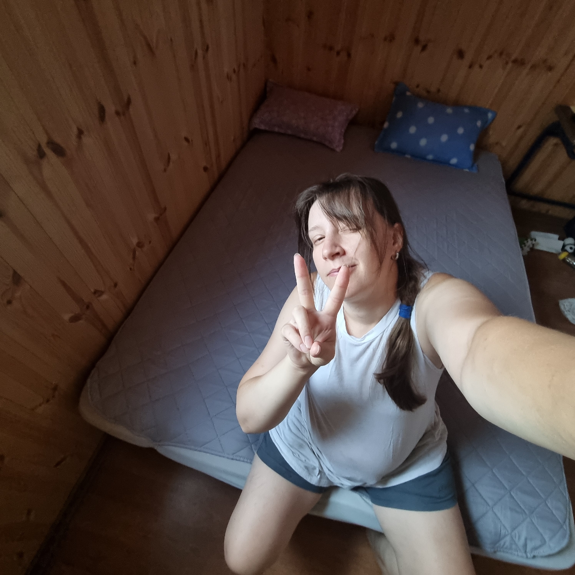 The image is a high-angle selfie taken by a woman inside a room with wooden walls and ceiling. She is kneeling on a mattress that covers most of the floor space. The mattress is gray and quilted. There are two pillows on the mattress: one is reddish-pink and the other is blue with white polka dots. The woman is wearing a white tank top and dark shorts. She is making a peace sign with her right hand and smiling. The overall impression is that of a casual, informal setting, possibly a cabin or a rustic room. There is no discernible indication of the image being taken in Dunnae-myeon, South Korea.