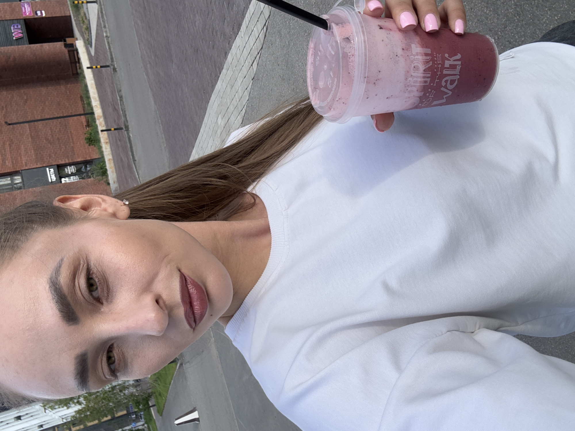 A medium-shot selfie shows a woman with brown hair pulled back in a ponytail, holding a pink berry smoothie in a clear plastic cup with a black straw. She is wearing a white crewneck sweatshirt and has on dark lipstick and light makeup. The woman is outdoors with a blurred background of a city street and brick buildings. There are bollards and a white traffic cone visible in the background. The text "SPIRIT FOR THE WALK" is visible on the cup.