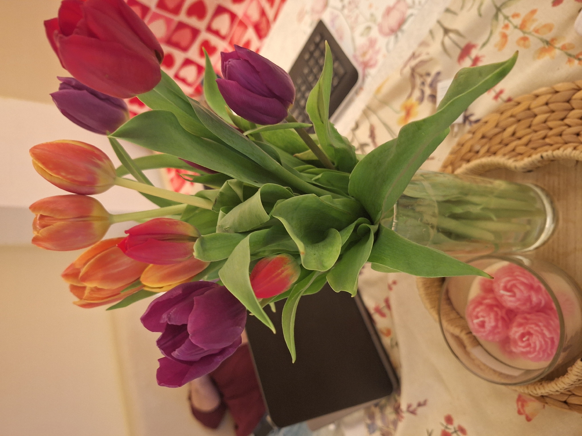 The image shows a bouquet of mixed tulips in a glass vase, placed on a table covered with a floral patterned tablecloth. The tulips are of varying colors, including red, purple, orange, and a multi-toned orange-red. There are numerous green leaves surrounding the flowers. Beside the vase, there is a laptop computer and a small, round woven basket containing what appear to be rose-shaped candles in a glass container. Part of a wall is visible in the background. The table also has a red and white patterned element. A pair of red shoes and feet are partially visible near the lower left corner, suggesting a person is seated nearby. The specific location in Varna, Bulgaria cannot be identified from the image alone.