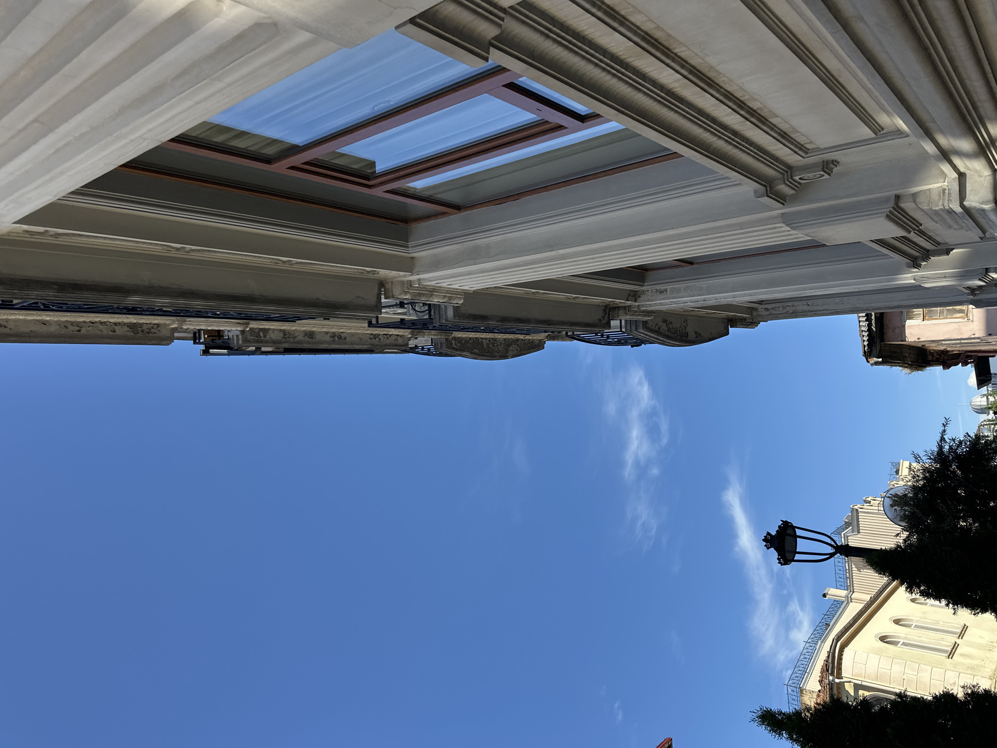 The image, taken from a ground-level perspective in Batumi, Georgia, showcases a view looking upwards towards the sky. The primary subjects are architectural elements and the sky itself. In the upper portion of the image, the ornate facade of a building, potentially an old structure, is visible. This includes elements like a skylight, decorative columns, and moldings, suggesting a historical or classic architectural style. The sky is a clear blue, with some streaks of clouds present. In the lower portion of the frame, parts of another building are visible, along with a street lamp and parts of a tree. The overall composition suggests an urban setting, with the buildings and sky being the main focus.