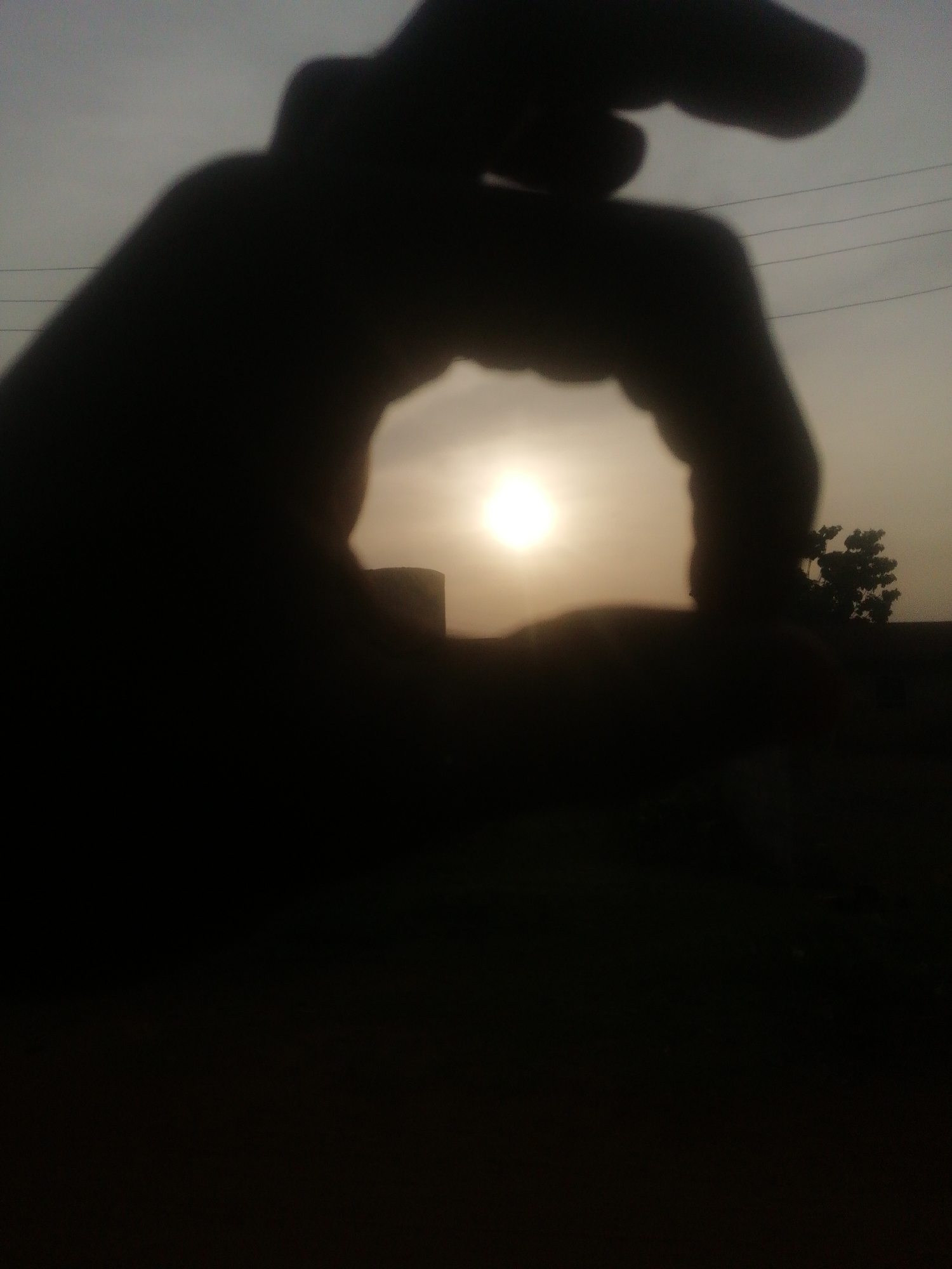 A dark silhouette of a hand with fingers curved into a circle frames a bright sun. The background includes electrical lines and a building or wall. A tree is visible on the right side of the frame.
