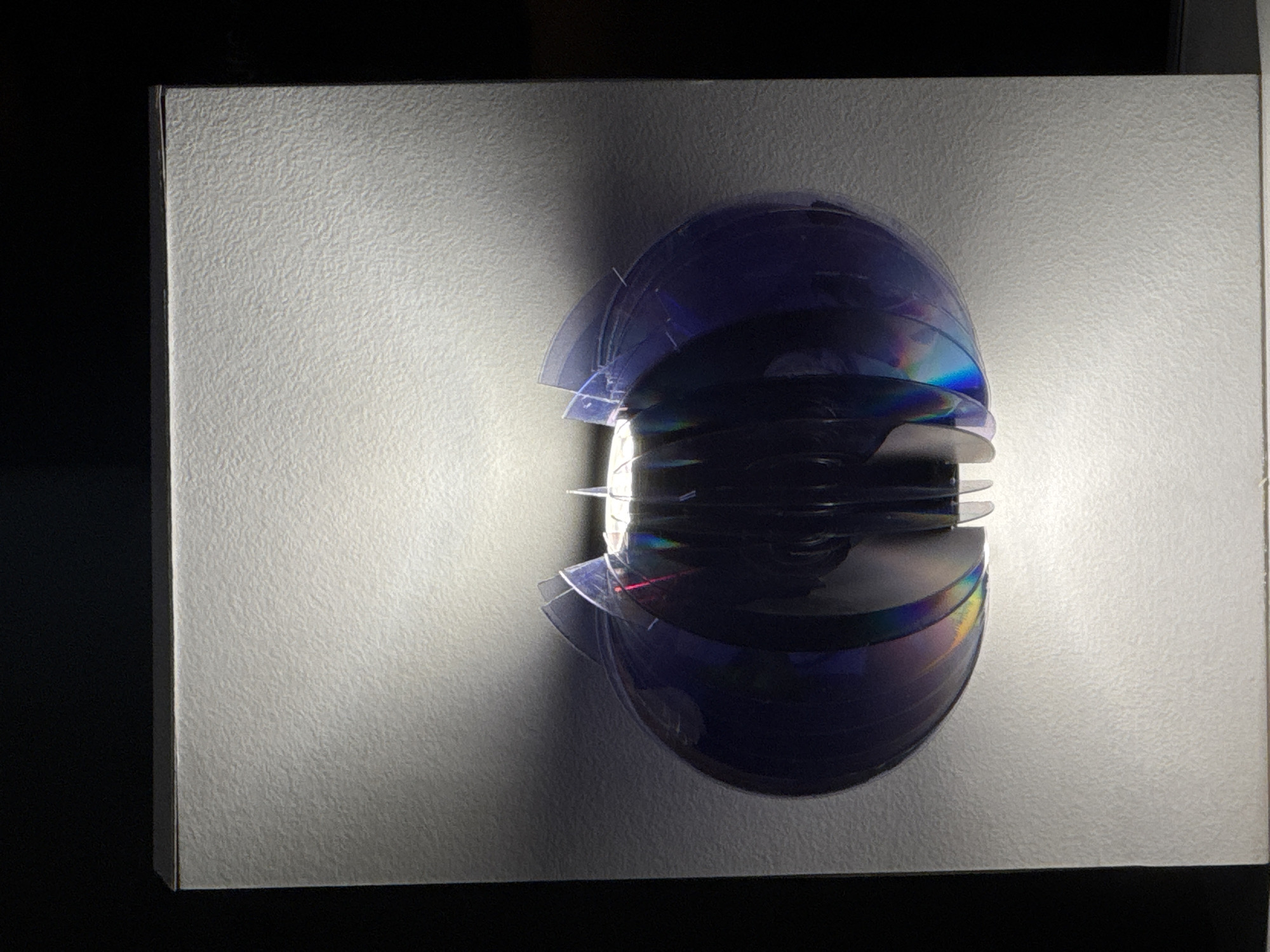 A still life photograph shows a stack of dark iridescent CDs or DVDs arranged in a vertical column at the center right of a rectangular, matte white surface. The light source from behind and to the right of the object casts a radial glow of light and shadow outwards onto the surface. The surface is resting on a dark, possibly black, background. The image does not contain any people or indications of a location in San Andrés Cholula, Mexico.