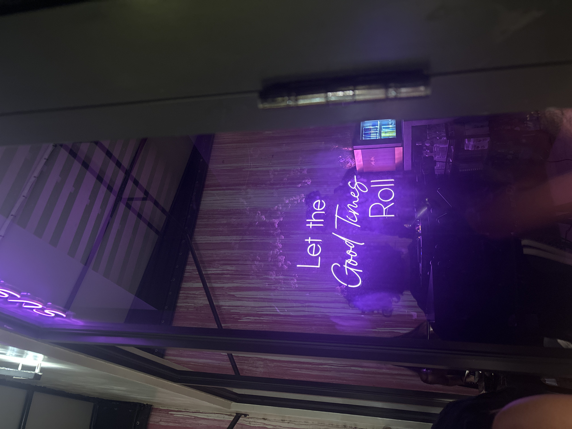 The image shows a neon sign in Miami, United States of America that says "Let the Good Times Roll." The sign appears to be inside a bar or restaurant, based on the background, but is partially obscured by the frame. The background is a wall with vertical stripes in shades of pink and white. Some items are visible on shelves in the background. The image appears to have been taken through a glass window or door.