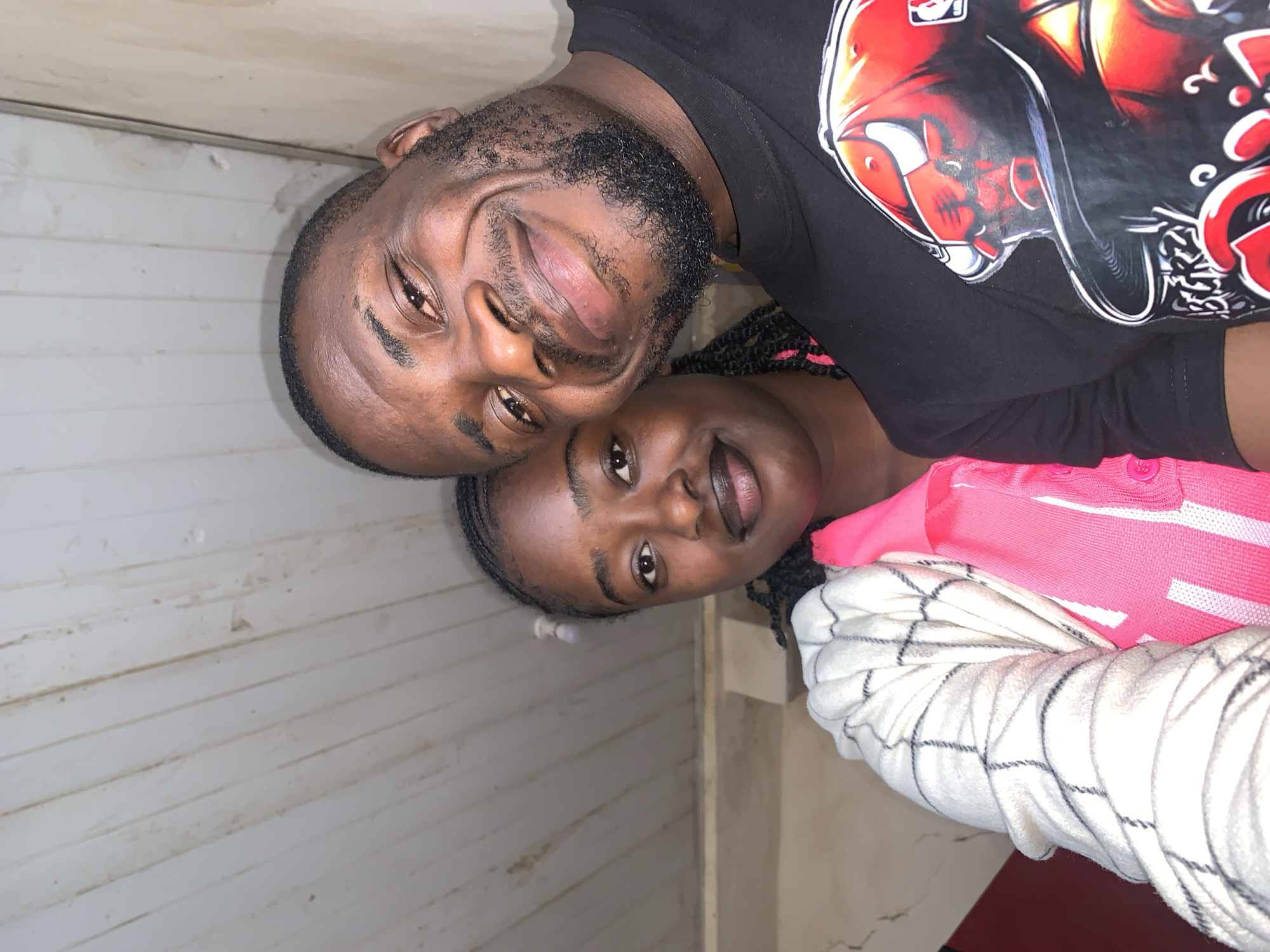 An eye-level selfie depicts two individuals, an adult male and an adult female, positioned in close proximity, likely indoors in New Stadium, Nigeria. The male, on the right, has dark skin, a short dark beard, and a broad smile, revealing teeth. He is wearing a black crew-neck t-shirt featuring a prominent red graphic, appearing to be a stylized bull or similar creature, with white accents. The female, on the left, also has dark skin and braided hair, exhibiting a subtle, closed-mouth smile. She wears a vibrant pink collared shirt beneath a white, long-sleeved outer garment with a dark grid or plaid pattern. The background consists of a light-colored, planked ceiling exhibiting minor discoloration and dirt, along with light-colored walls showing signs of wear and minor staining.