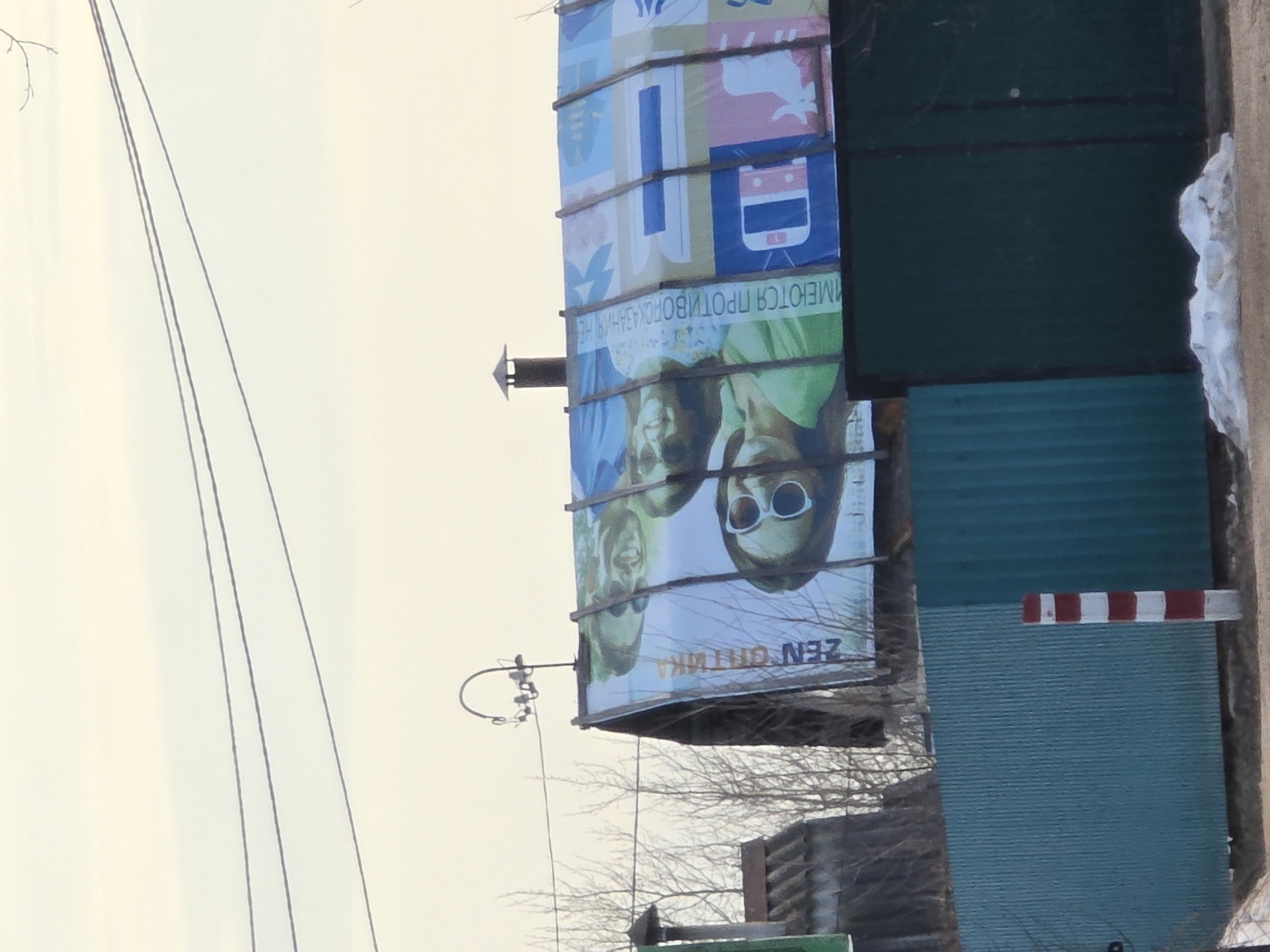The image depicts a vertical billboard in Noviy, Russia. The billboard displays an advertisement featuring two individuals, one wearing sunglasses. Above the figures is text in Russian. The top of the billboard has a colorful, patterned design that includes an image of a train. The billboard is situated near a green corrugated metal structure, possibly a fence or building, with a red and white striped pole protruding from it. Power lines are visible against the sky in the background. The surrounding environment includes bare tree branches and what appears to be a rooftop.