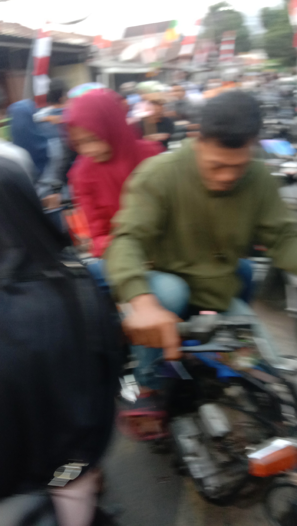 In Pesantren, Indonesia, a group of people are riding motorcycles in a congested area with buildings and trees in the background. The image shows two individuals on the foreground motorcycle. The driver wears a green long-sleeved shirt and light blue jeans, while the passenger in front of him wears a red hooded jacket. A third person, seen from the back and wearing a black garment, is on another motorcycle to the left of the frame. The scene is blurred, suggesting motion, and the lighting indicates it is daytime. The presence of Indonesian flags in the background hints at a local event or celebration.
